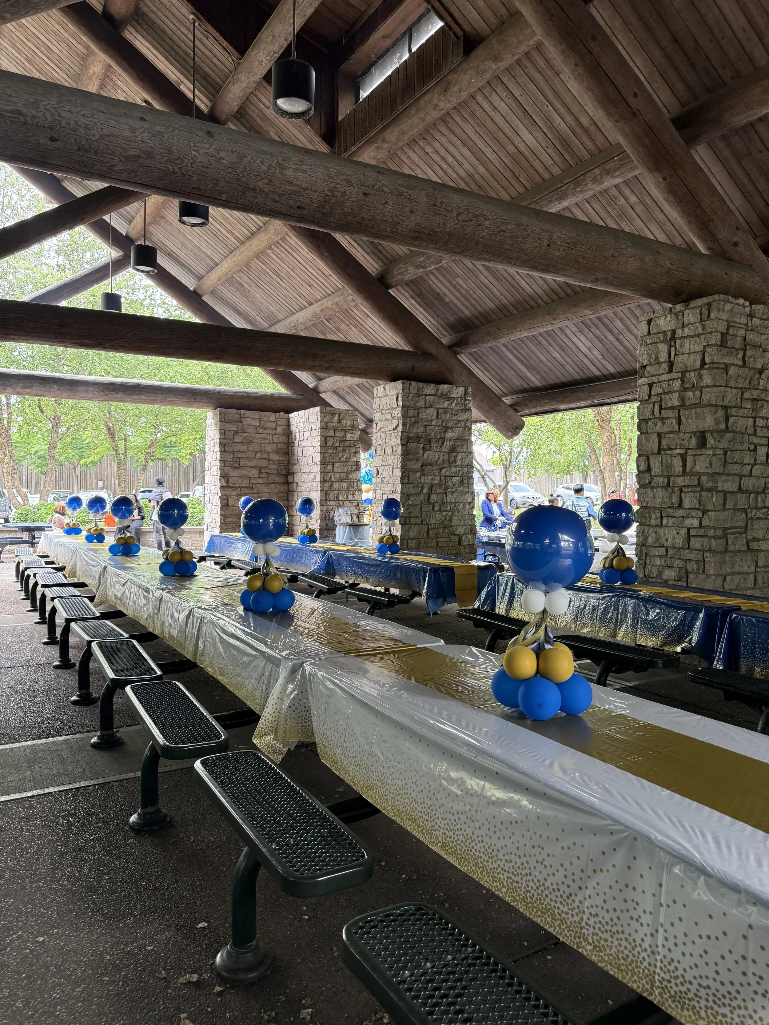 Indoor shelter with decorated tables and balloons for a celebration, with outdoor trees and people visible in the background.