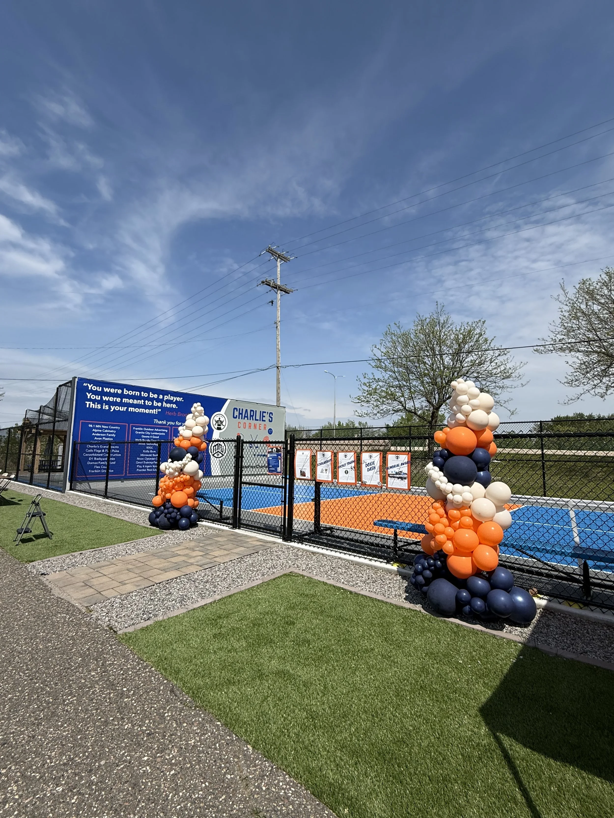 Outdoor sports court surrounded by a black fence, with balloon decorations on either side, and a blue sign with white text. Trees and a utility pole are visible in the background under a partly cloudy sky.