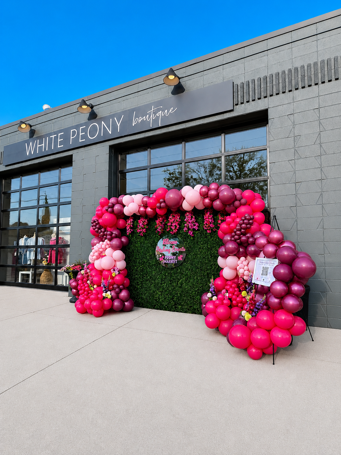 Storefront with a large pink and purple balloon arch and a sign reading 'White Peony Boutique'. The store has large glass windows with clothing displayed inside, and the sky is clear and blue.