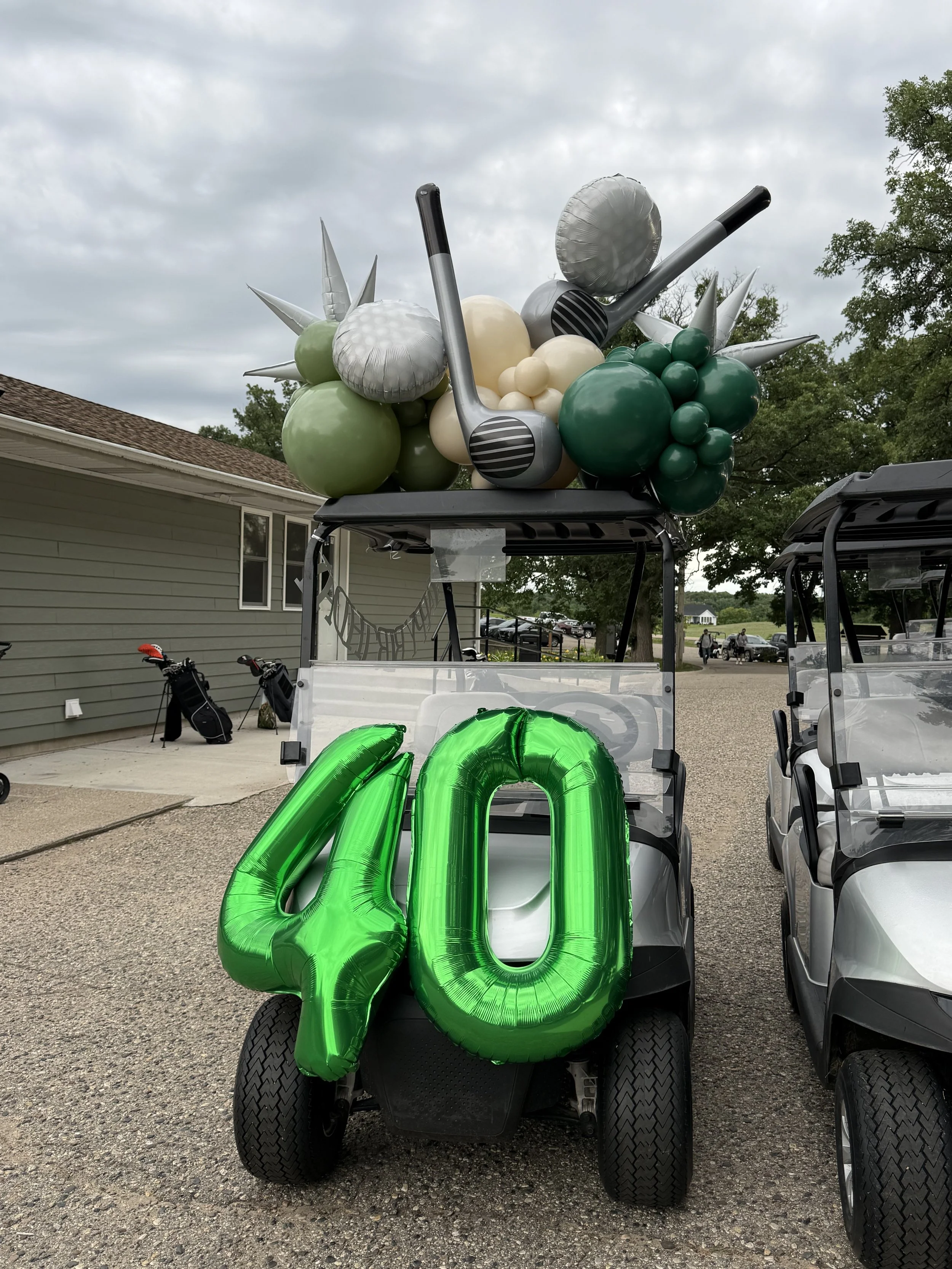 Golf cart decorated with green and silver balloons and large green foil balloon numbers '20'. There are golf clubs in the background.