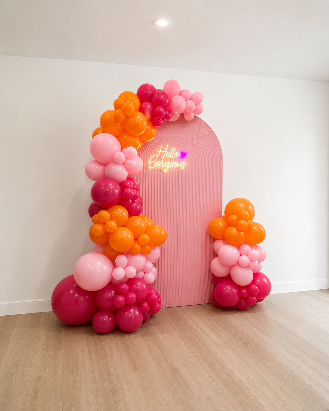Decorative balloon arch with pink, orange, and red balloons surrounding a pink backdrop with a neon sign that says 'Hello Gorgeous' in cursive with a purple heart, on a wooden floor with white walls.