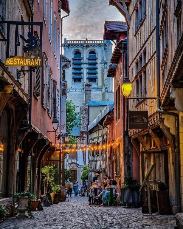 Troyes - Ruelle pavée avec des bâtiments en pierre, des lampadaires, des potted plants, des gens assis à une terrasse de restaurant, et un grand bâtiment en arrière-plan.