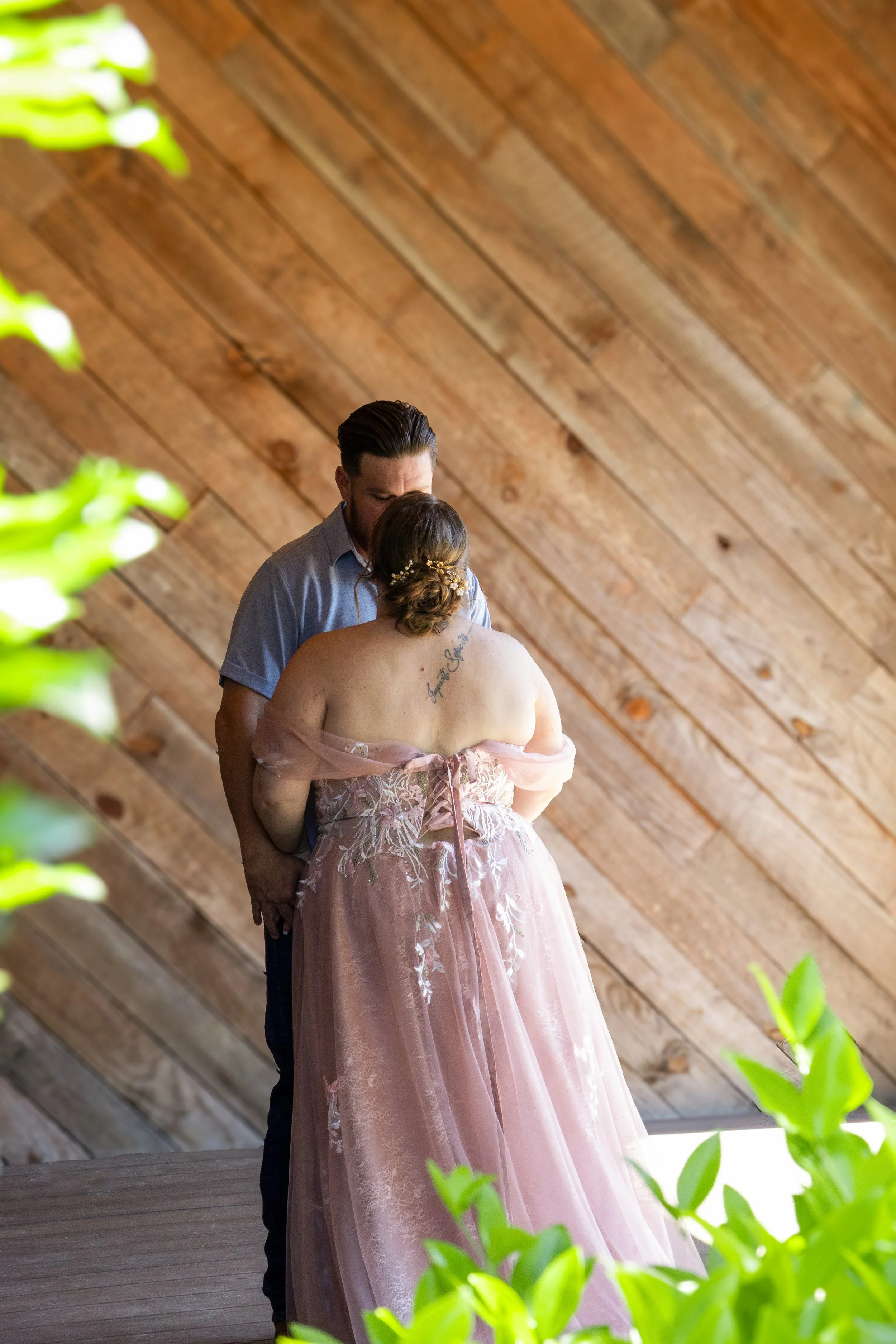 A couple sharing a kiss during their wedding ceremony inside a venue with a wooden ceiling. The bride is wearing a pink off-shoulder dress with lace details and a floral hairpiece, and the groom is dressed in a light blue shirt.