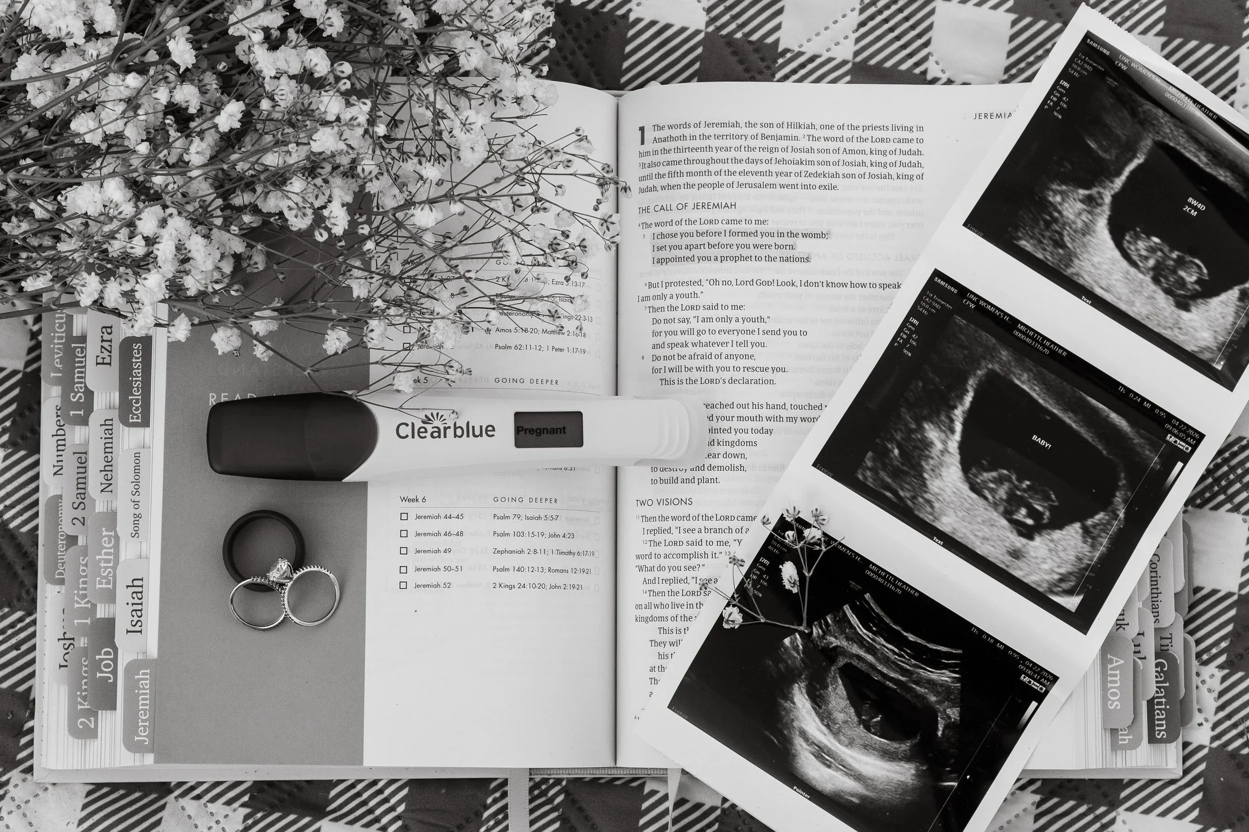 A flat lay arrangement featuring a Bible open to Jeremiah, alongside ultrasound images, a pregnancy test labeled 'Pregnant' from Clearblue, a wedding ring, a pair of rings, a flower bouquet, a book titled 'Ecclesiastical' and other books, all on a patterned cloth.