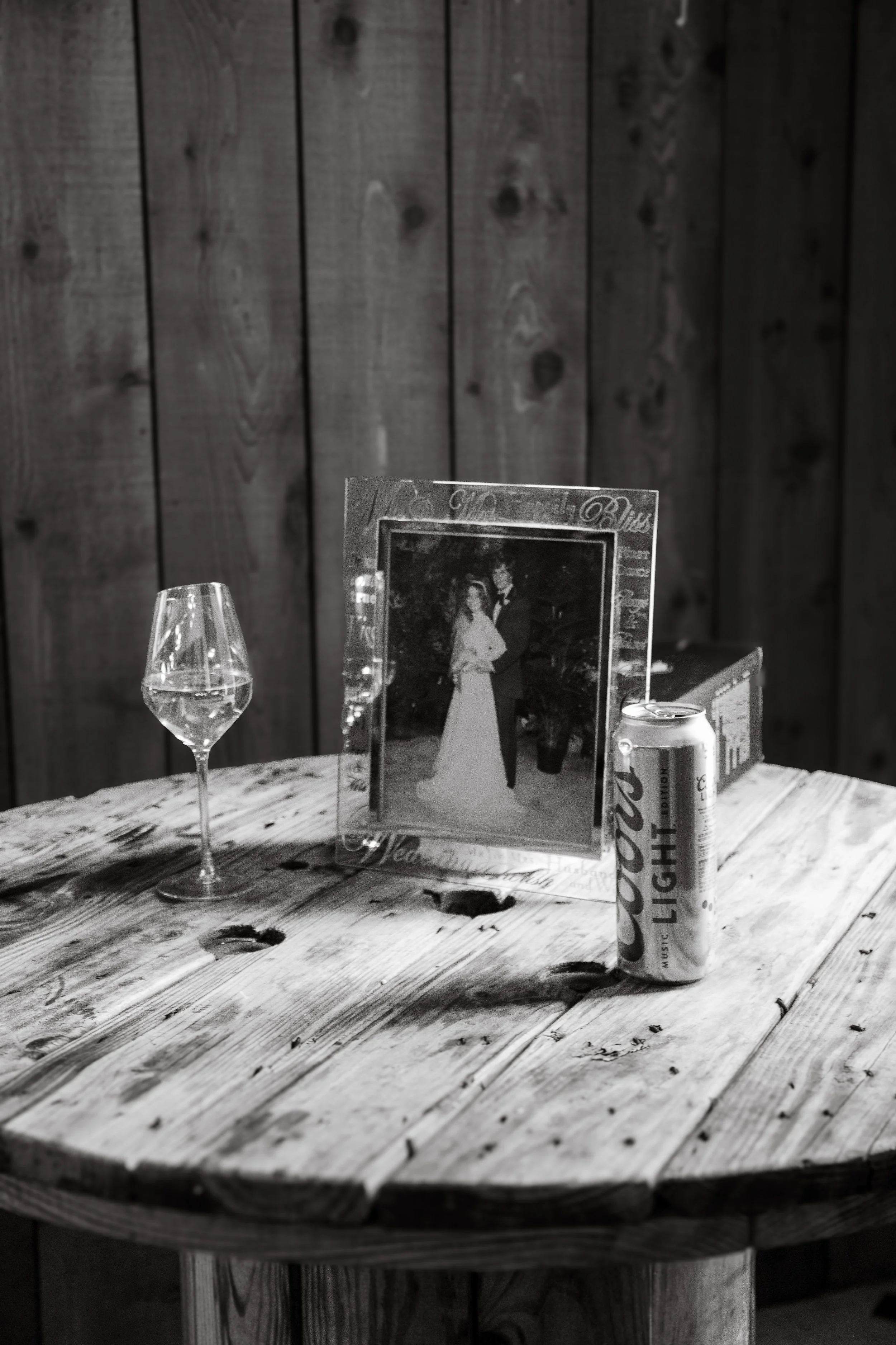 A wooden table with a wine glass, a can of Coors Light beer, and a framed photograph of a couple in wedding attire. The background features dark wooden paneling.