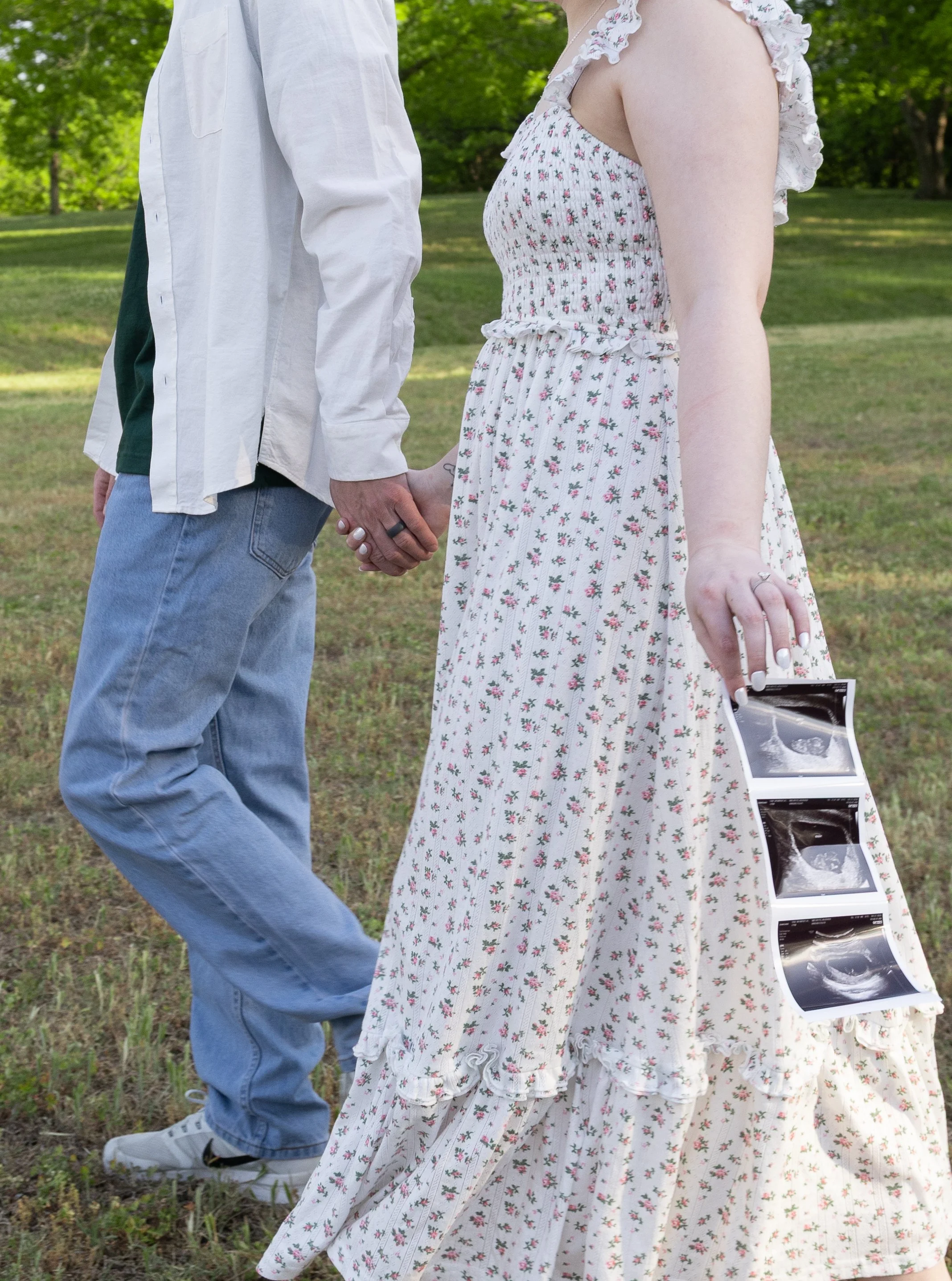 A person wearing a floral maxi dress holding ultrasound images standing next to another person in casual clothing, outdoors on grass with trees in the background.
