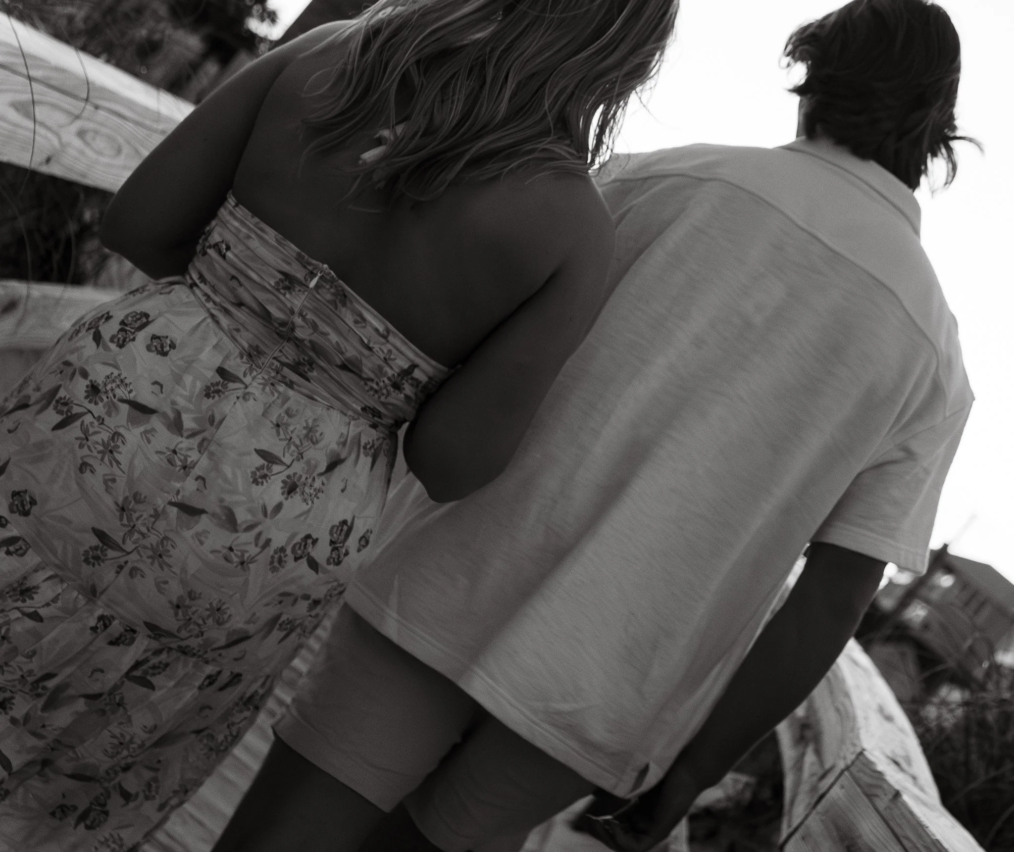 A black and white photograph of a woman sitting on a boat or dock, looking away from the camera. She has shoulder-length wavy hair and is wearing a sleeveless floral dress. There is a man seated next to her, partially visible, wearing a light-colored, button-up shirt.