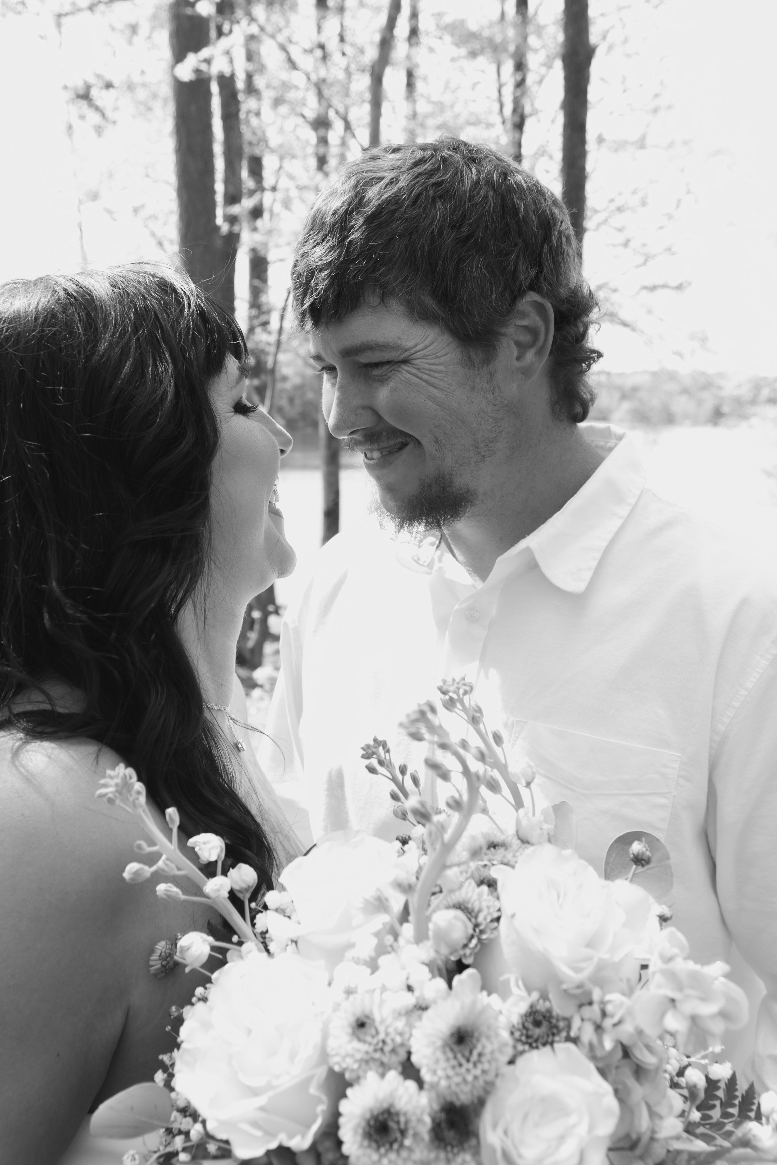 A black-and-white photograph of a smiling couple facing each other closely outdoors, with trees in the background, and flowers in the foreground.