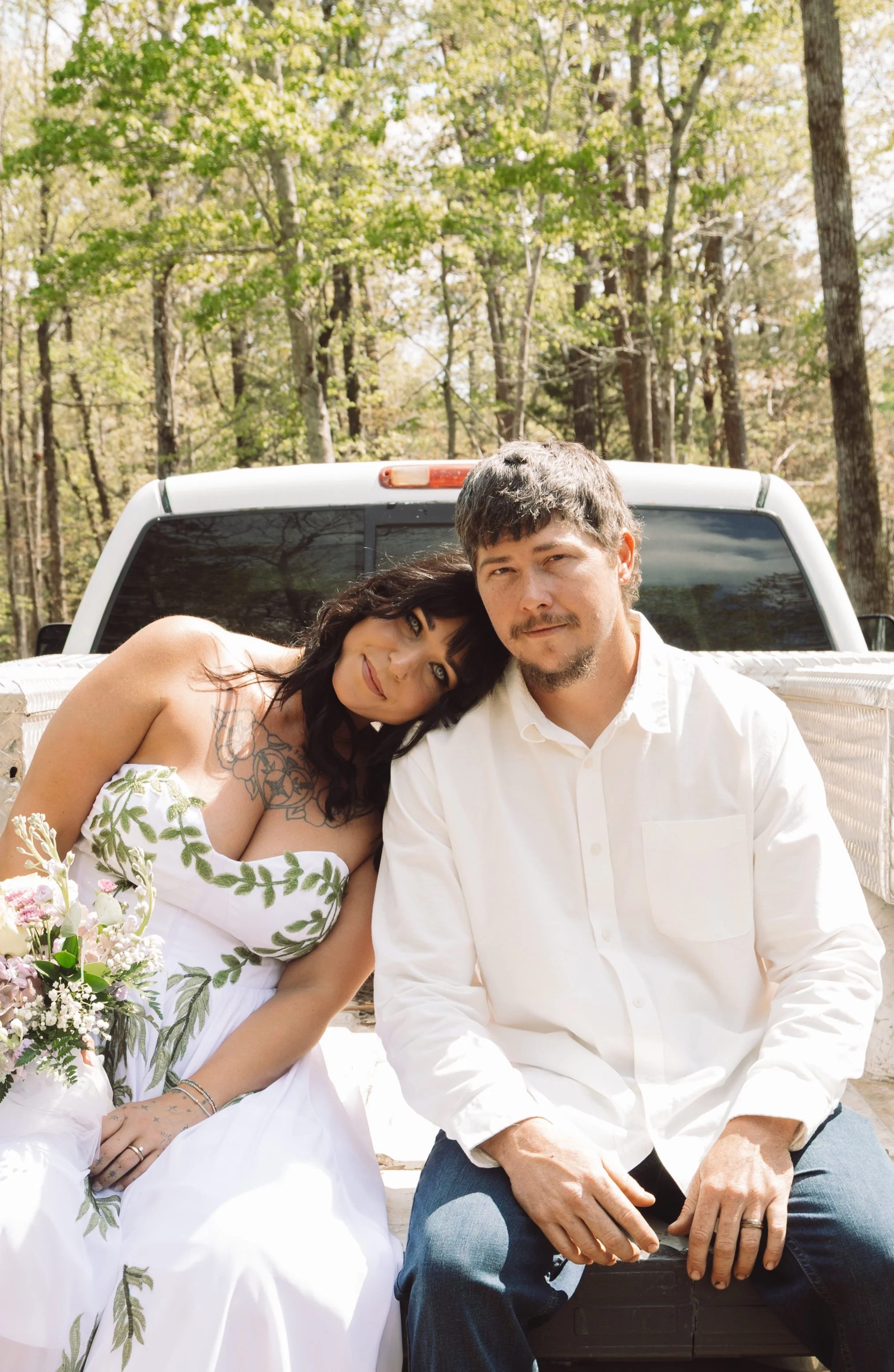 A man and woman are sitting closely together outdoors, with the woman resting her head on the man's shoulder, both smiling softly at the camera. They are seated on the back of a pickup truck with a background of trees and greenery, suggesting a forest or park setting.
