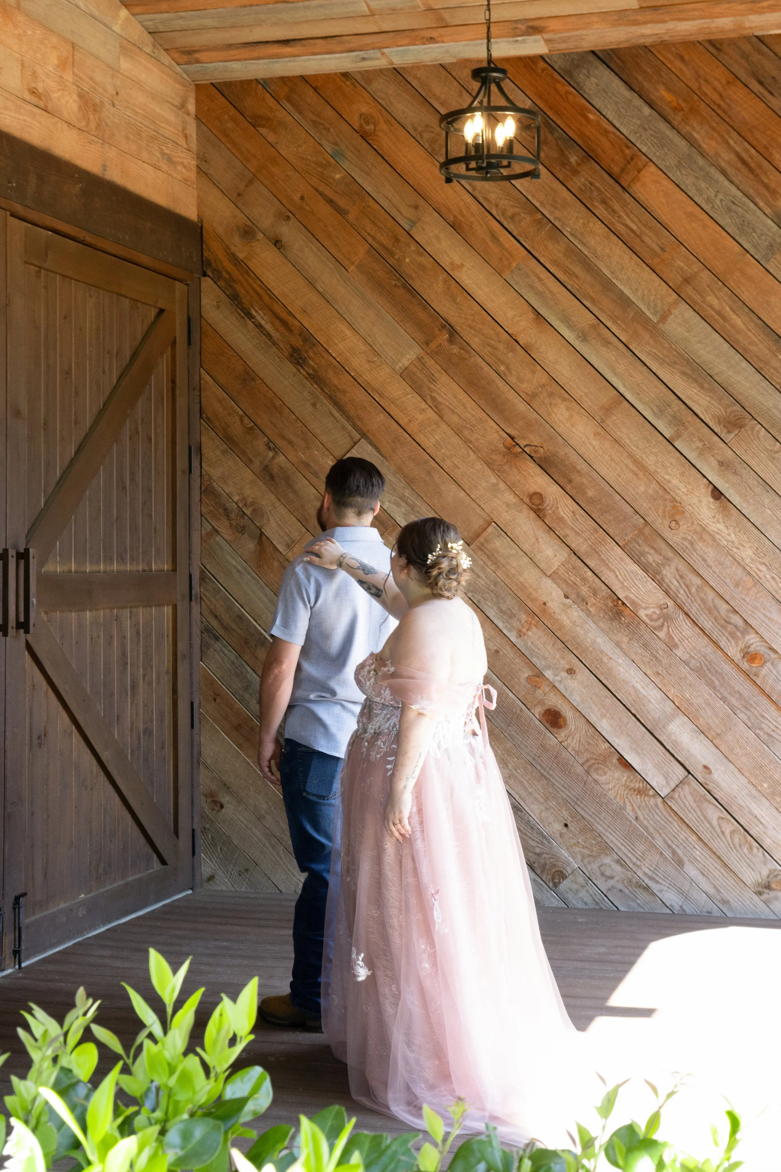 A couple, the woman in a pink gown and the man in a casual shirt, standing together in a rustic wooden room, possibly during a wedding or special event, with greenery in the foreground and a wooden wall with a hanging light fixture in the background.