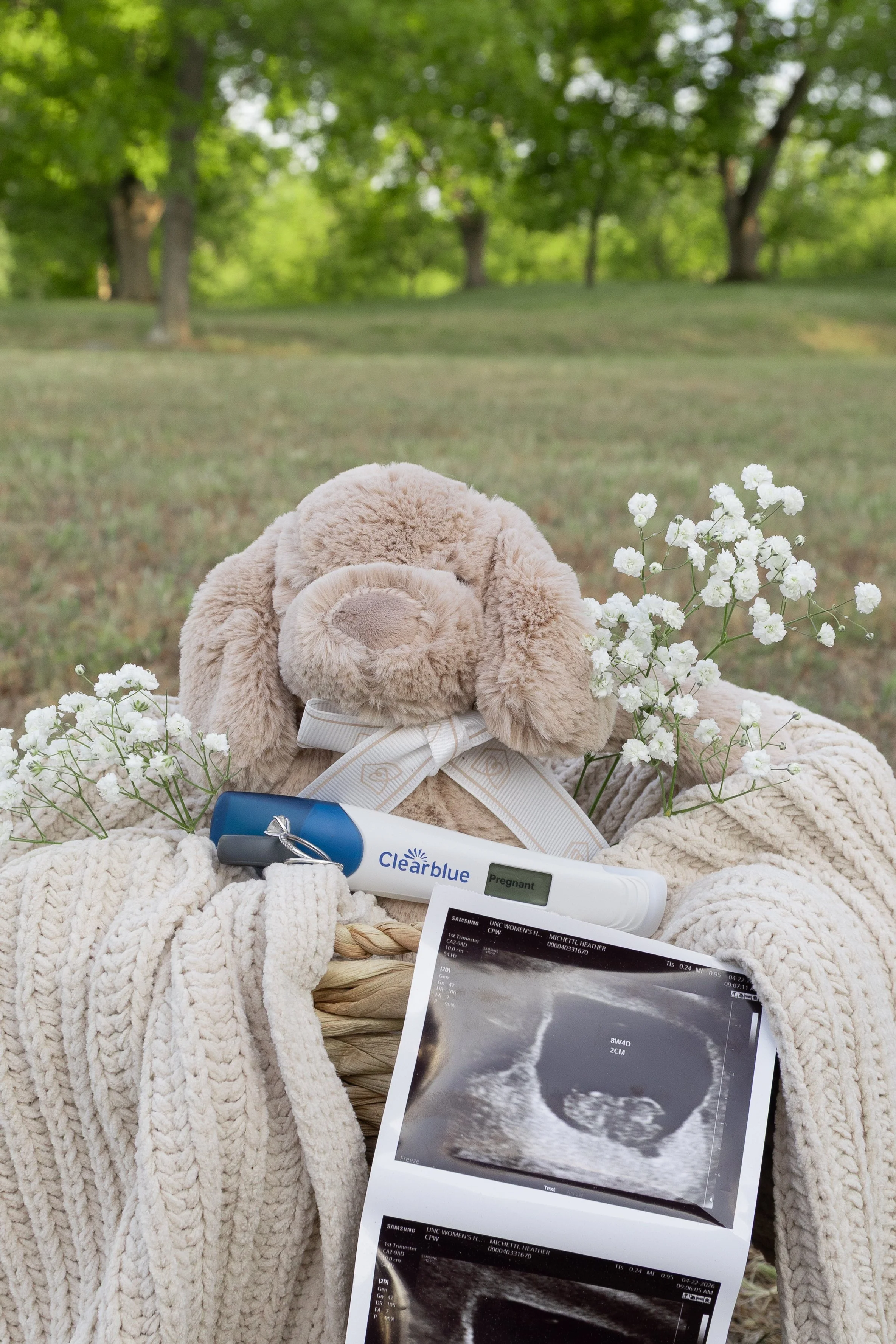 A plush teddy bear wearing a ribbon necktie, a bouquet of white flowers, a 3D ultrasound printout, a pregnancy test showing positive, and a set of keys placed on a thick white blanket outdoors. The background features a grassy area with trees.