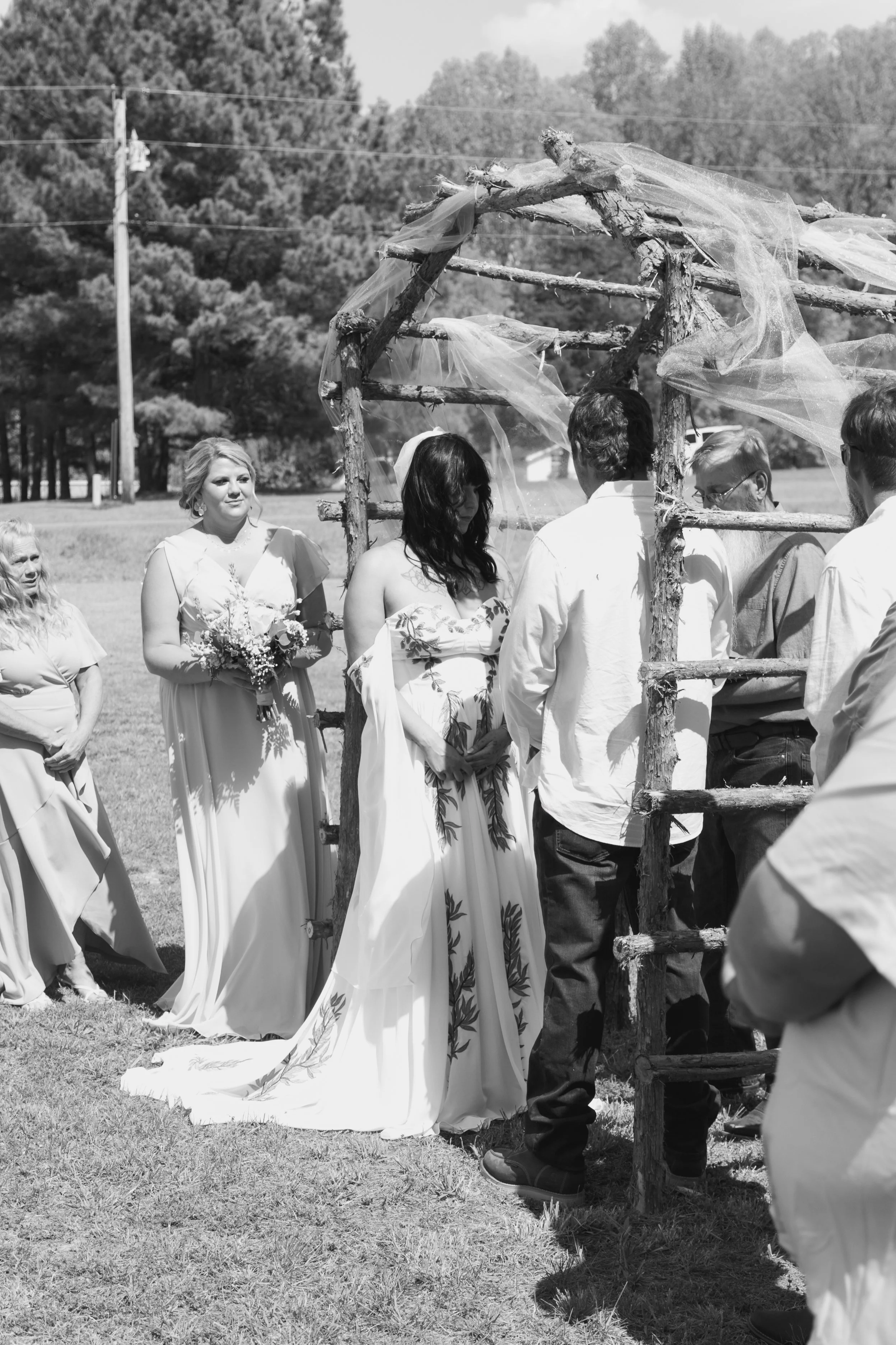 A group of people, including a woman in a wedding dress, gather outdoors under a rustic wooden arch decorated with tulle fabric, during a wedding ceremony on a sunny day with trees and utility poles in the background.