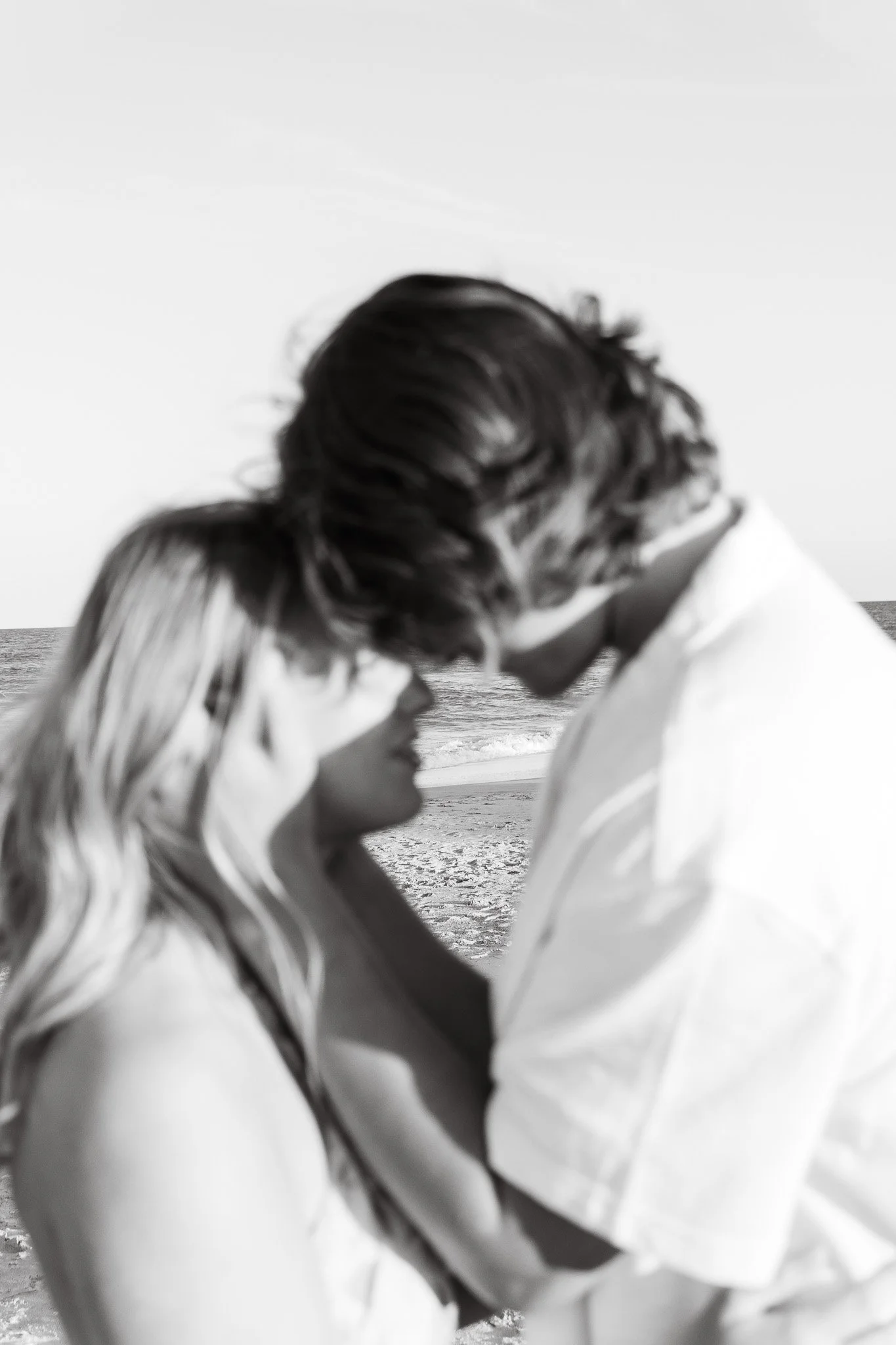 A black-and-white photograph of a couple on the beach, kissing, with ocean waves in the background.