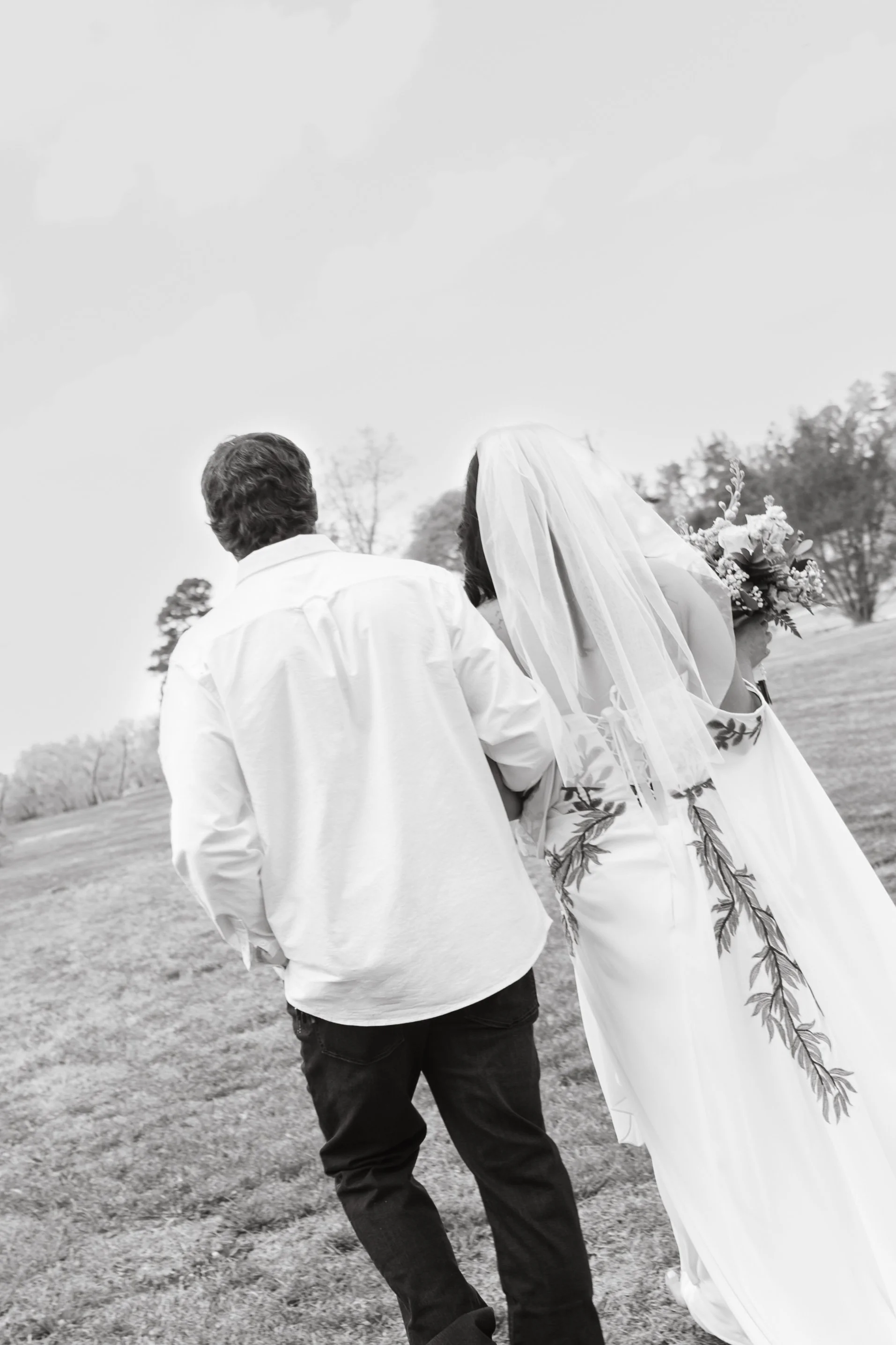 A black and white photo of a couple walking away outdoors, with the woman wearing a veil and holding a bouquet, and the man wearing a white shirt and dark pants.