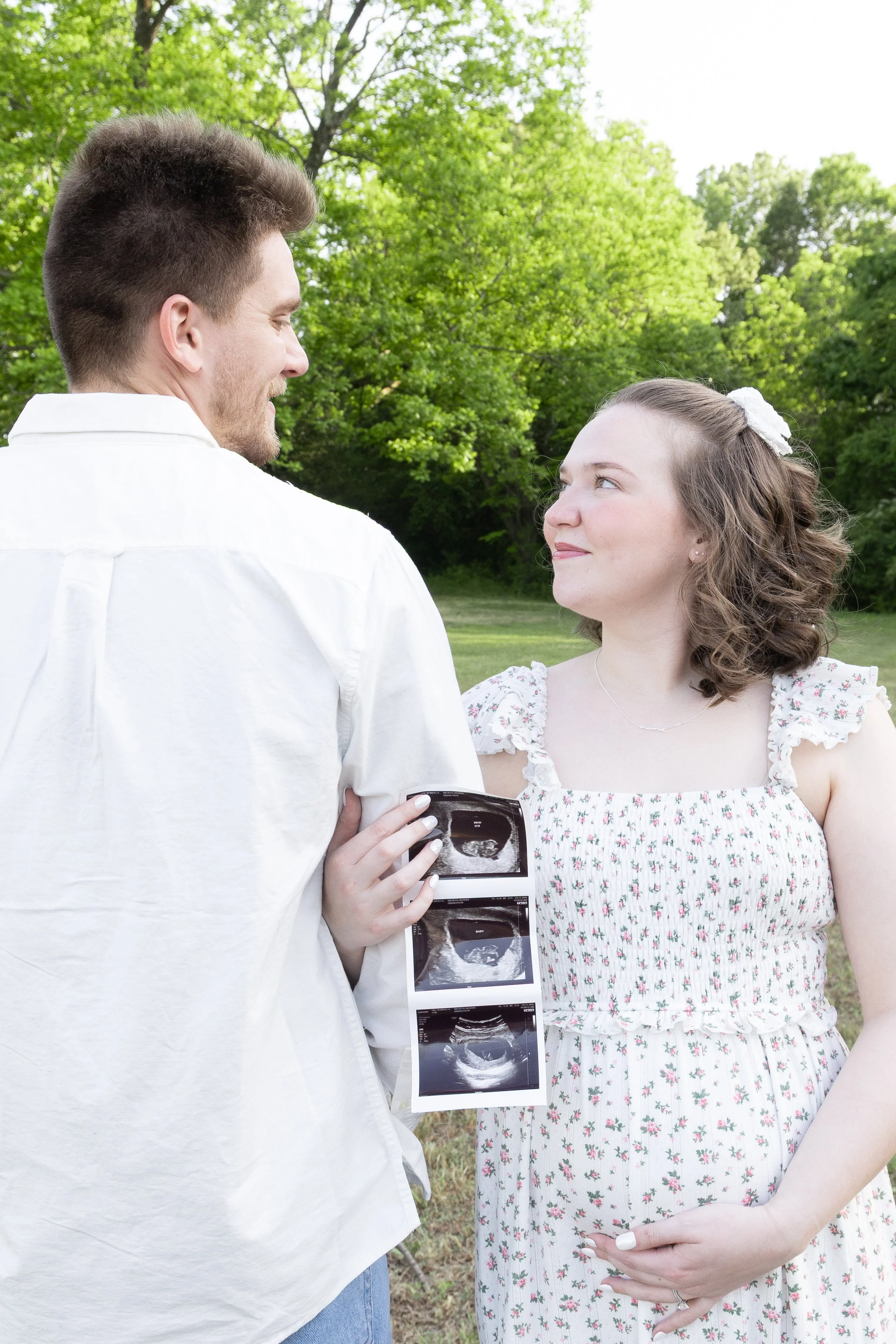 A man and woman are outdoors; the man is holding ultrasound images and they are looking at each other, smiling, during daytime with greenery in the background.