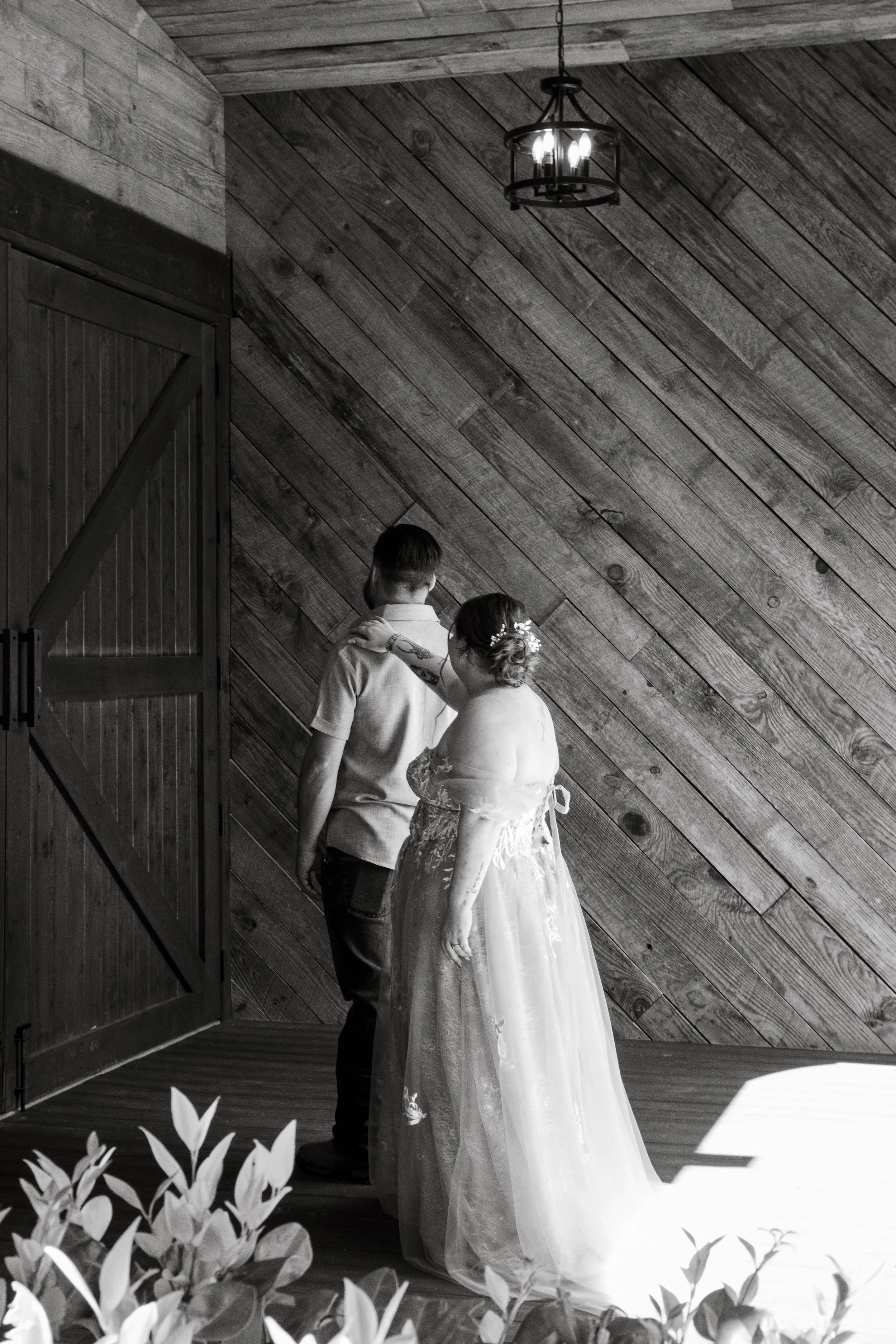 A bride and groom stand close together in a rustic indoor setting with wooden walls and ceiling. The bride wears a white wedding gown with lace details, and her hair is styled with small flowers. The groom wears a light-colored shirt and dark trousers. The bride's hand rests on the groom's shoulder. In the foreground, there are blurred plants, and a vintage chandelier hangs from the ceiling.