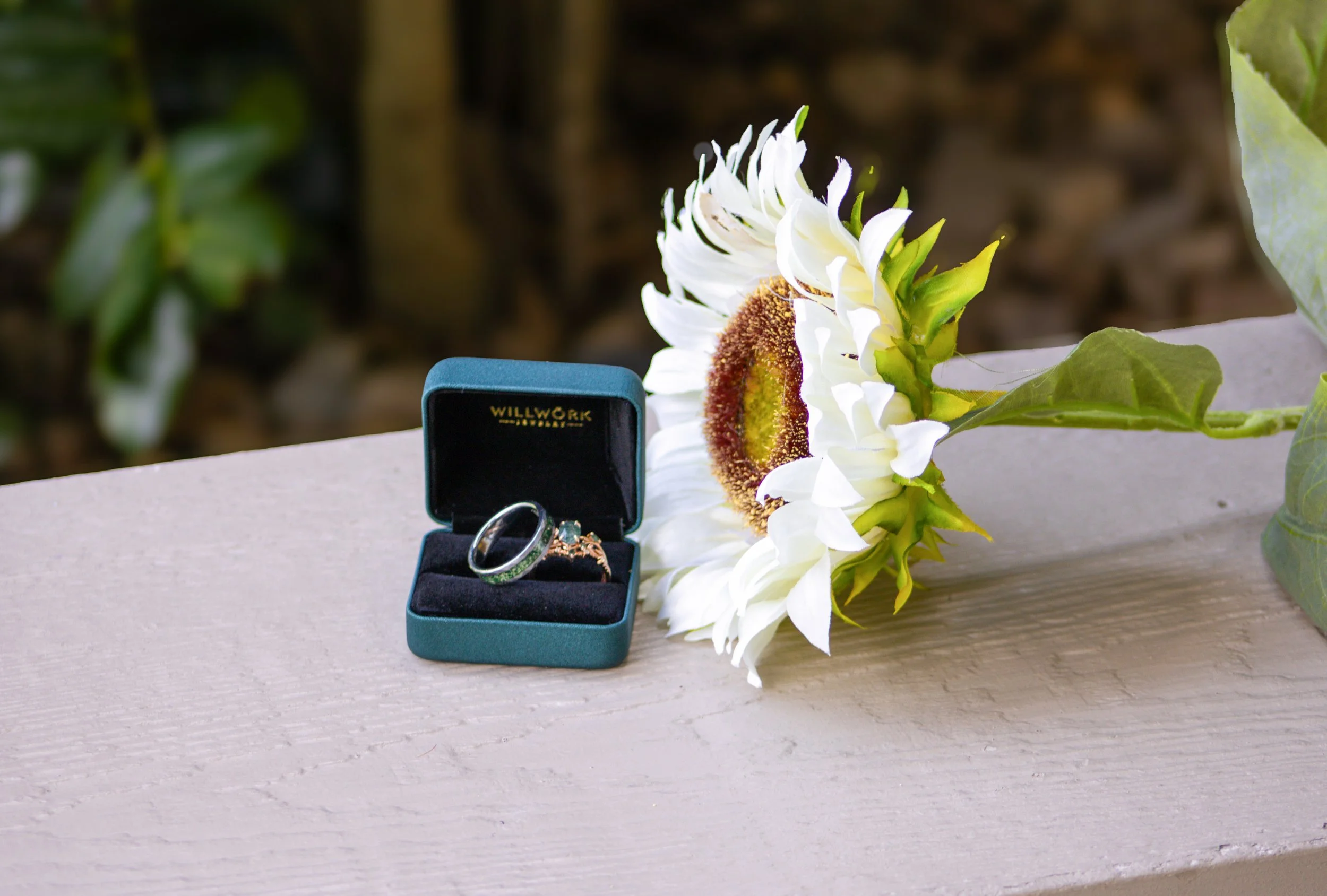 A pair of rings, one with a plain band and the other with intricate designs, inside a black box labeled 'Willwork Jewelry,' positioned on a light-colored wooden surface. A white flower with a brown and yellow center and green leaves lies next to the box. The background features blurred greenery and soil.