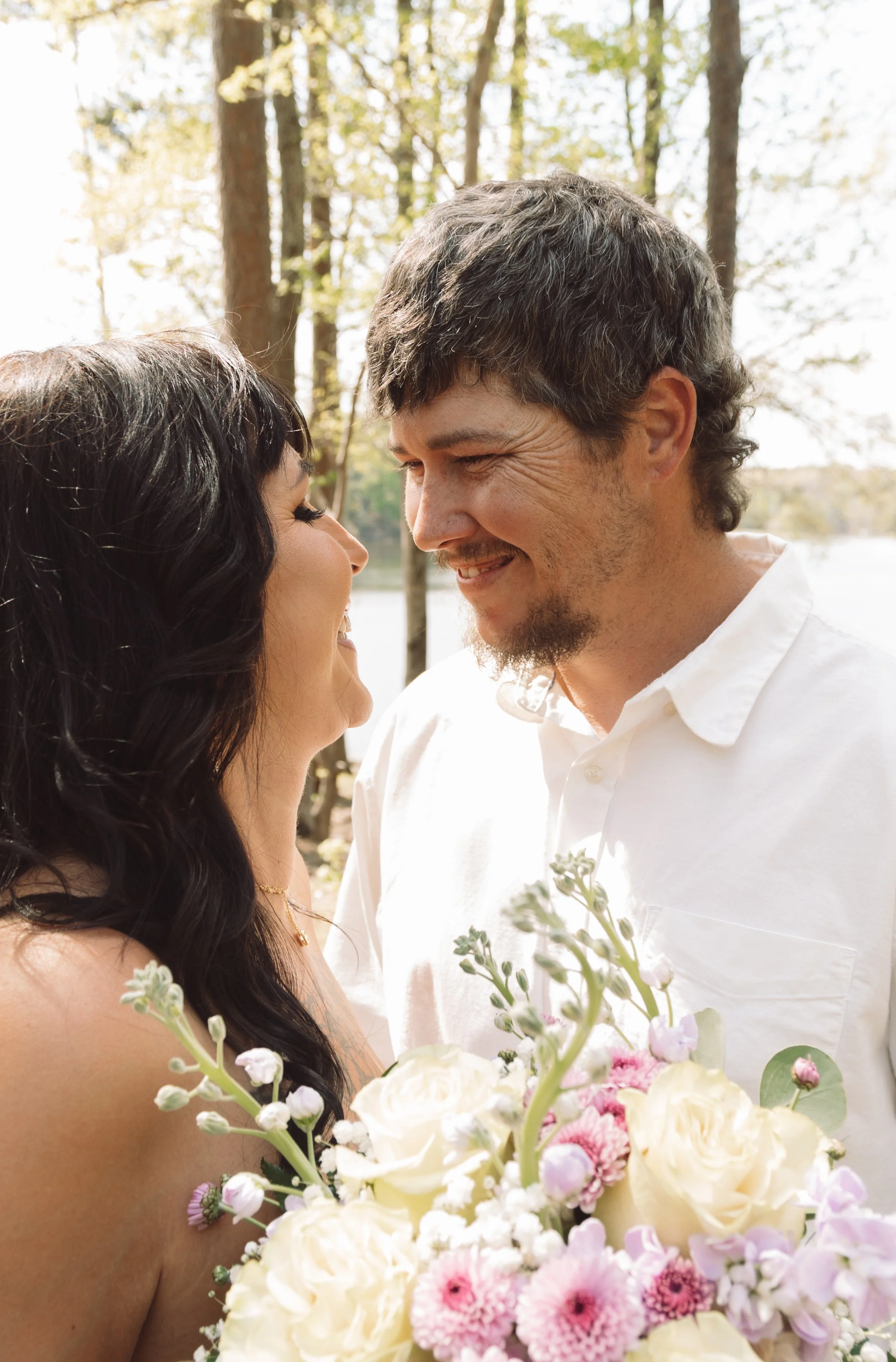 A couple stands close together outdoors, smiling and gazing into each other's eyes. The woman has long dark hair and is holding a bouquet of flowers. The man has short dark hair and a beard, wearing a white shirt. The background features trees and bright, natural daylight.