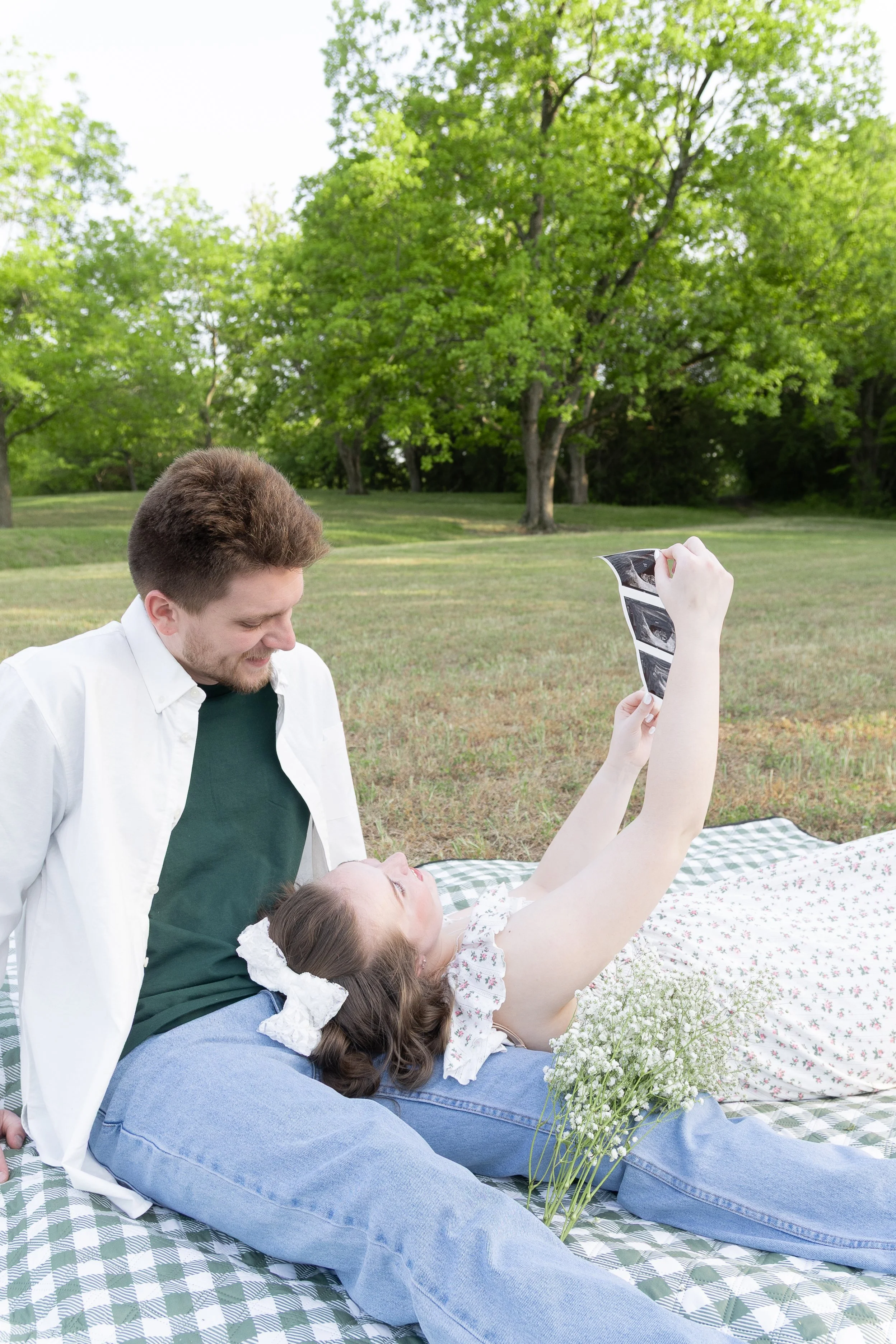 A family relaxing outdoors on a grassy field with trees in the background. A young girl lies on her back, holding up an ultrasound photo. A man and girl are smiling, enjoying the moment.