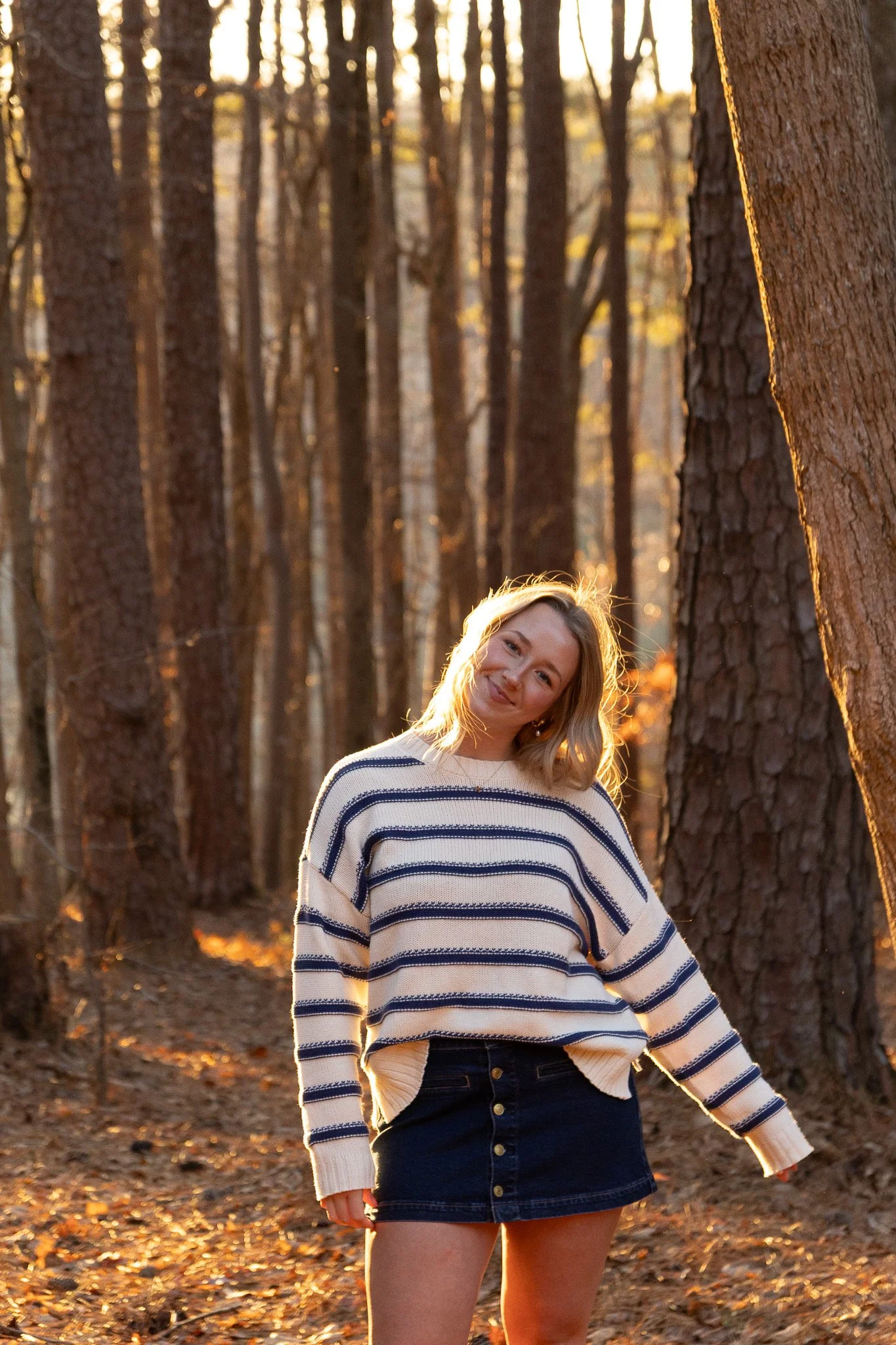 A woman smiling in a striped sweater and denim skirt standing in a forest with tall trees during sunset.