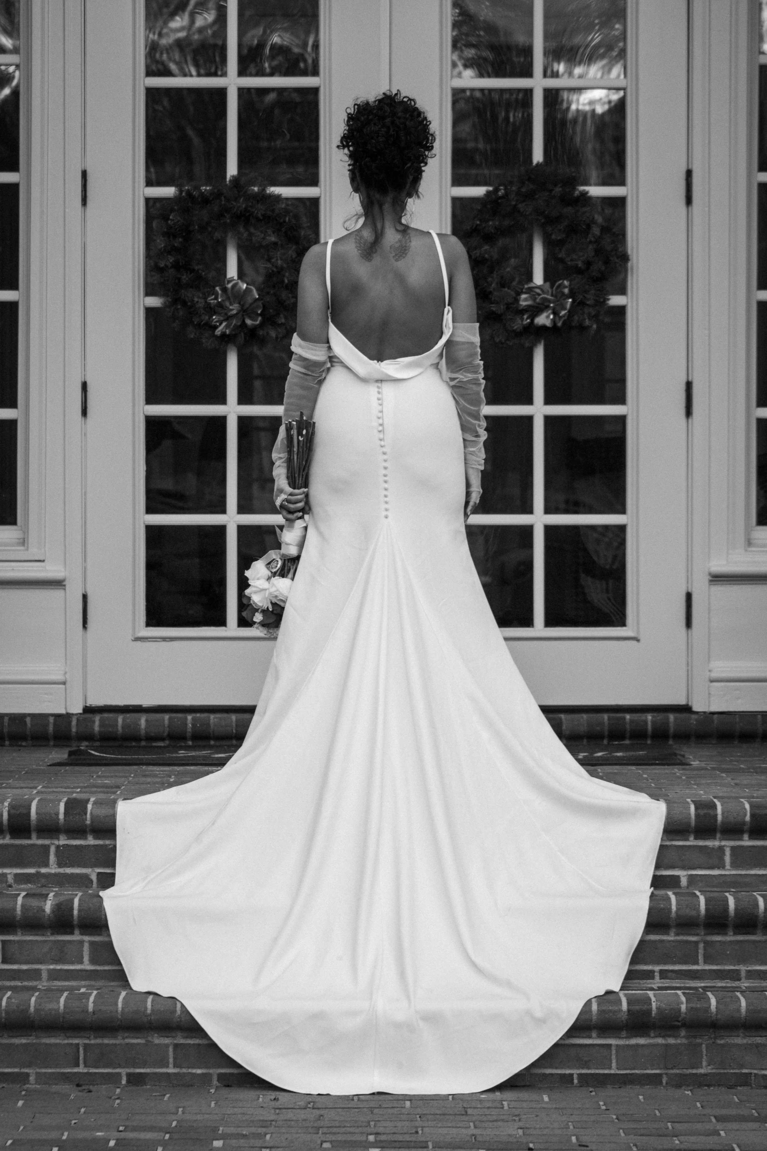 Back view of a woman in a wedding dress standing on brick steps in front of a glass door decorated with Christmas wreaths.
