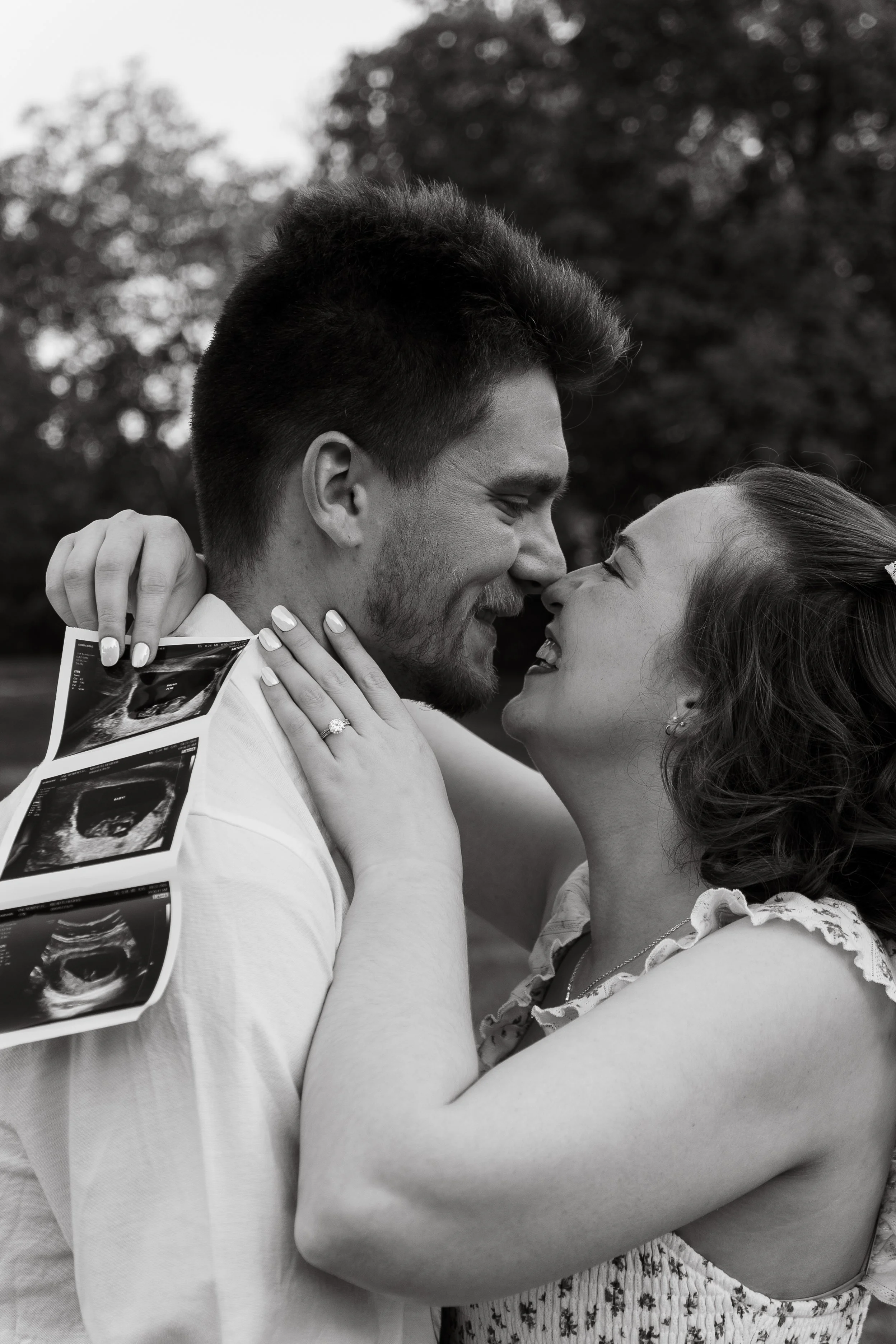 Black and white photo of a happy man and woman touching their noses together, with the woman holding ultrasound images and wearing an engagement ring.