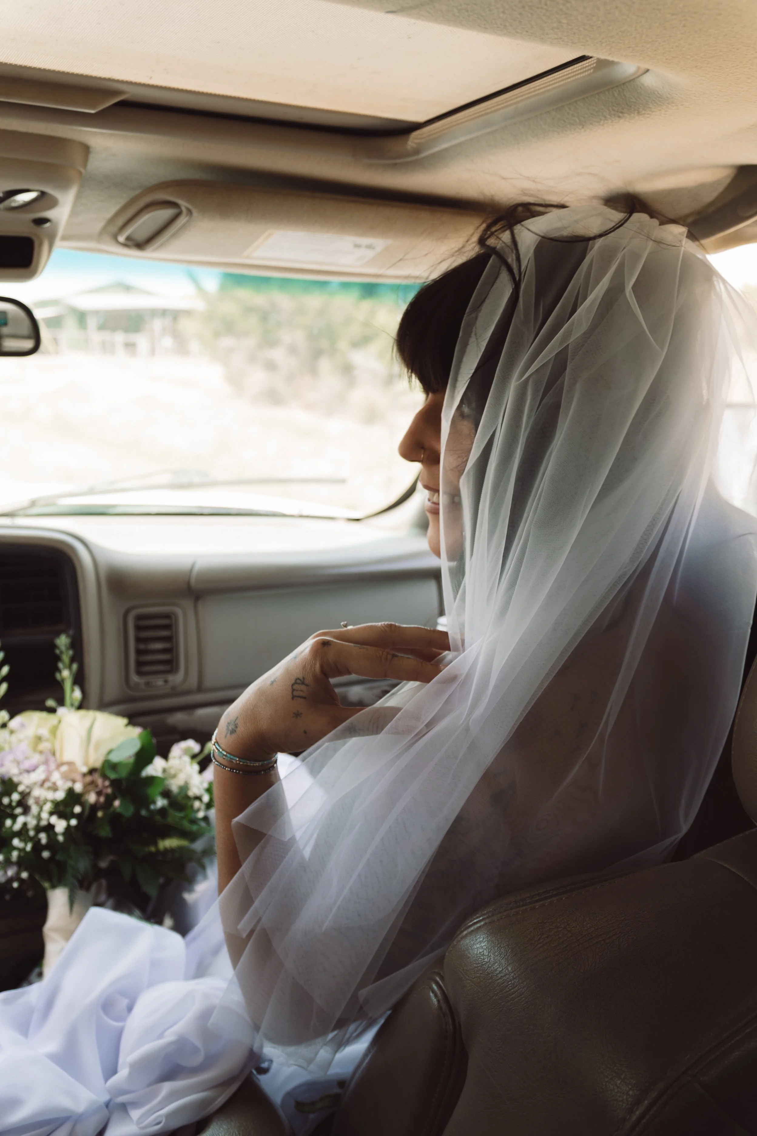 A woman in a wedding dress with a veil seated in a vehicle, with a wedding bouquet nearby.