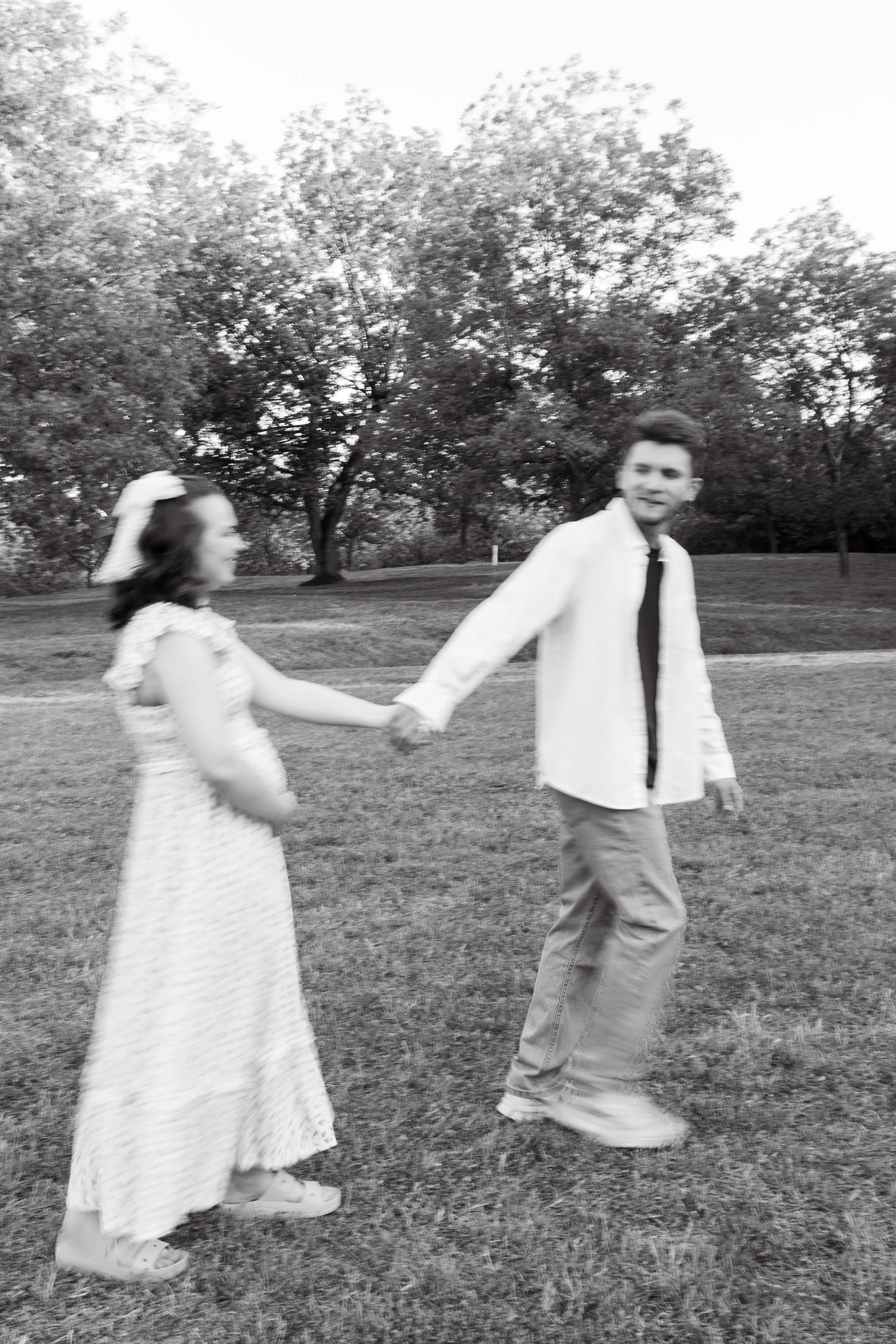 A black and white photo of a young man and a young girl holding hands and walking outdoors in a park with trees in the background.