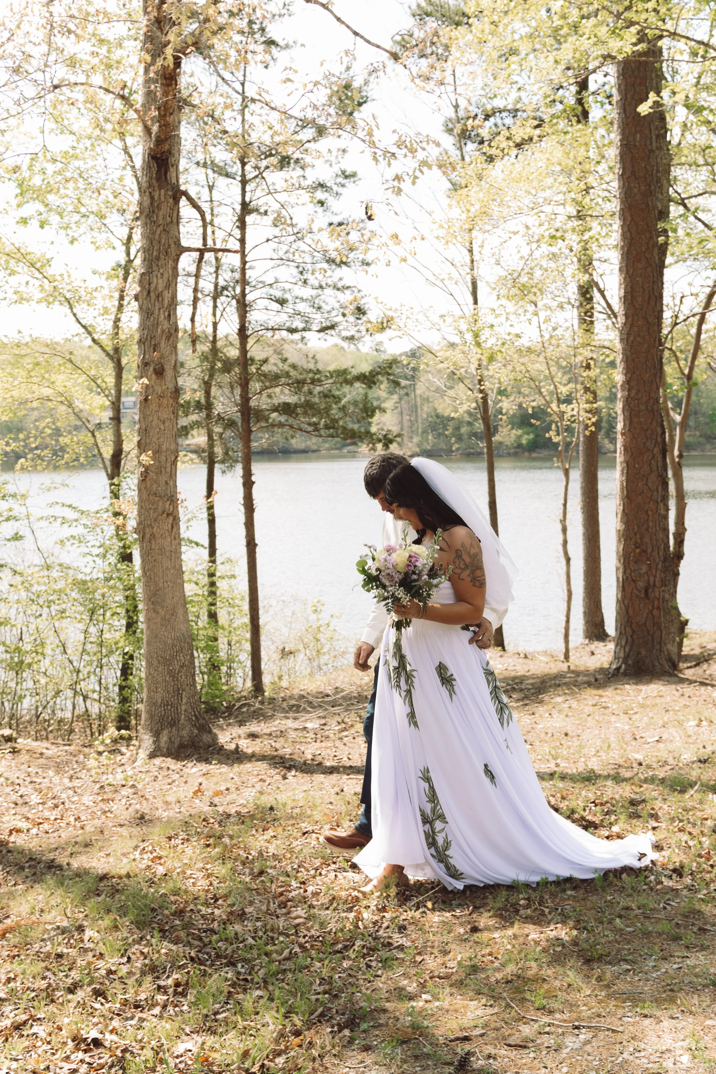 A bride with long dark hair and a white wedding dress with green leaf patterns, holding a bouquet, walking under trees near a lake. The groom is partially visible, dressed in dark pants and brown shoes, standing behind her.