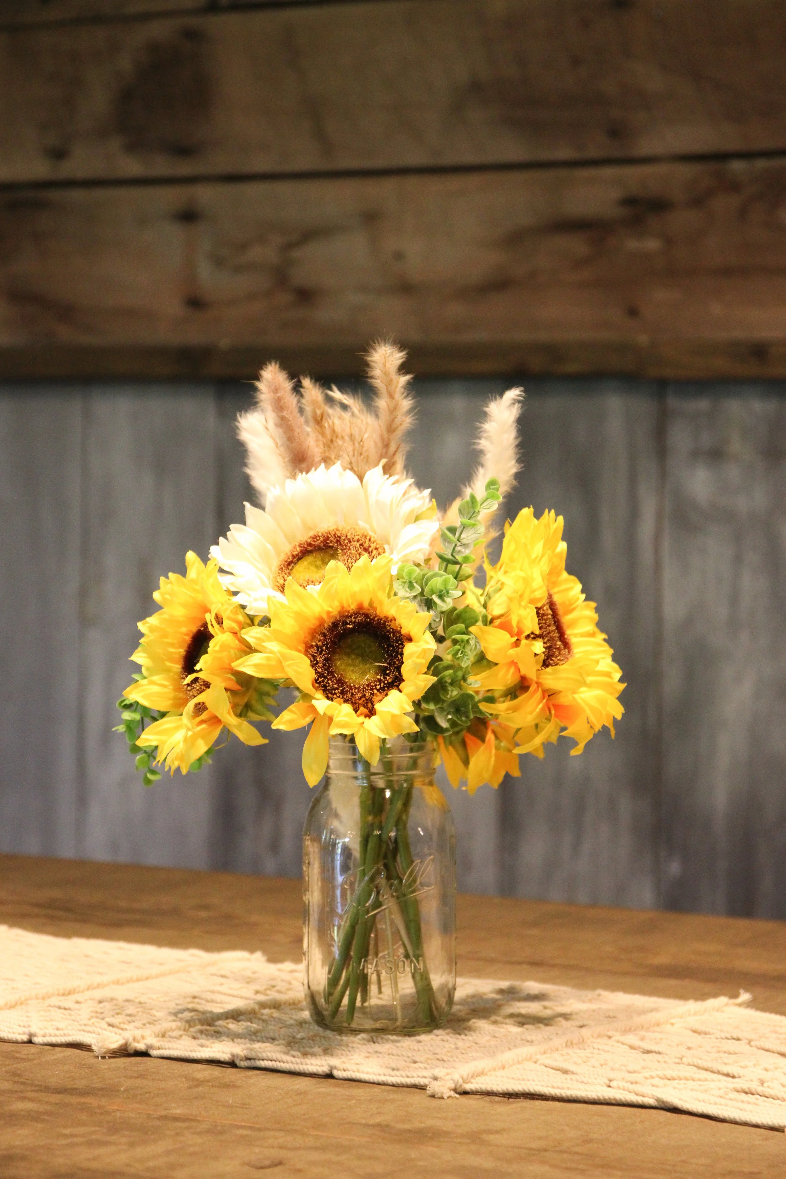 A bouquet of yellow sunflowers, white daisies, and greenery in a clear glass mason jar on a wooden table, with a textured beige cloth underneath and a gray wooden wall in the background.