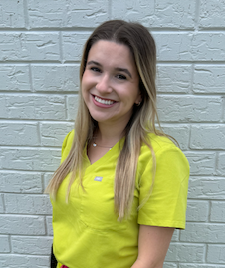 Young woman with long blonde hair, smiling, wearing a bright yellow shirt, standing against a white brick wall.