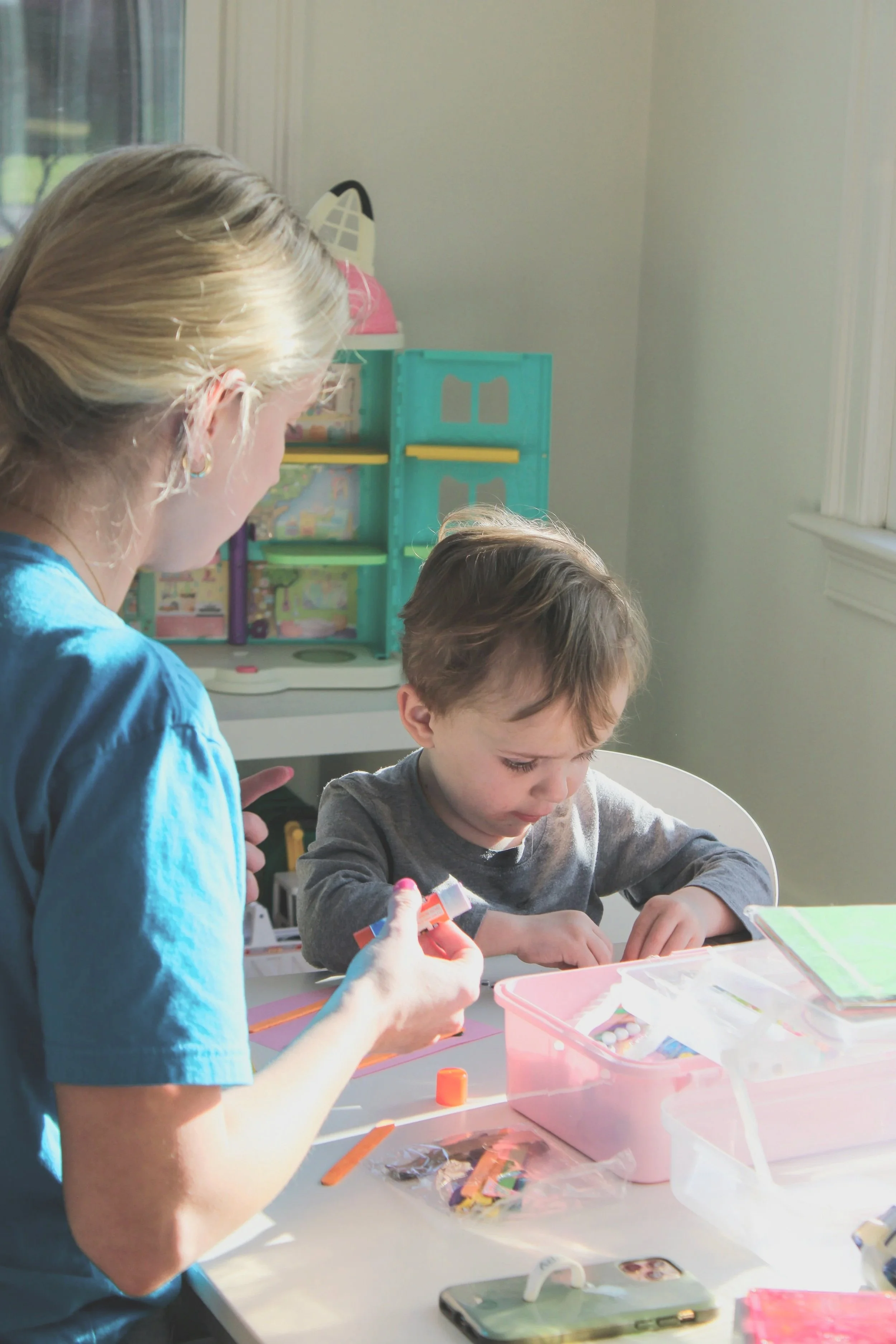 A woman and a young boy sit at a table working on arts and crafts. The woman is on the left with blonde hair and wearing a blue shirt, and the boy is on the right with brown hair in a gray shirt. There are various craft supplies on the table, including a pink box, colored paper, and small craft items.