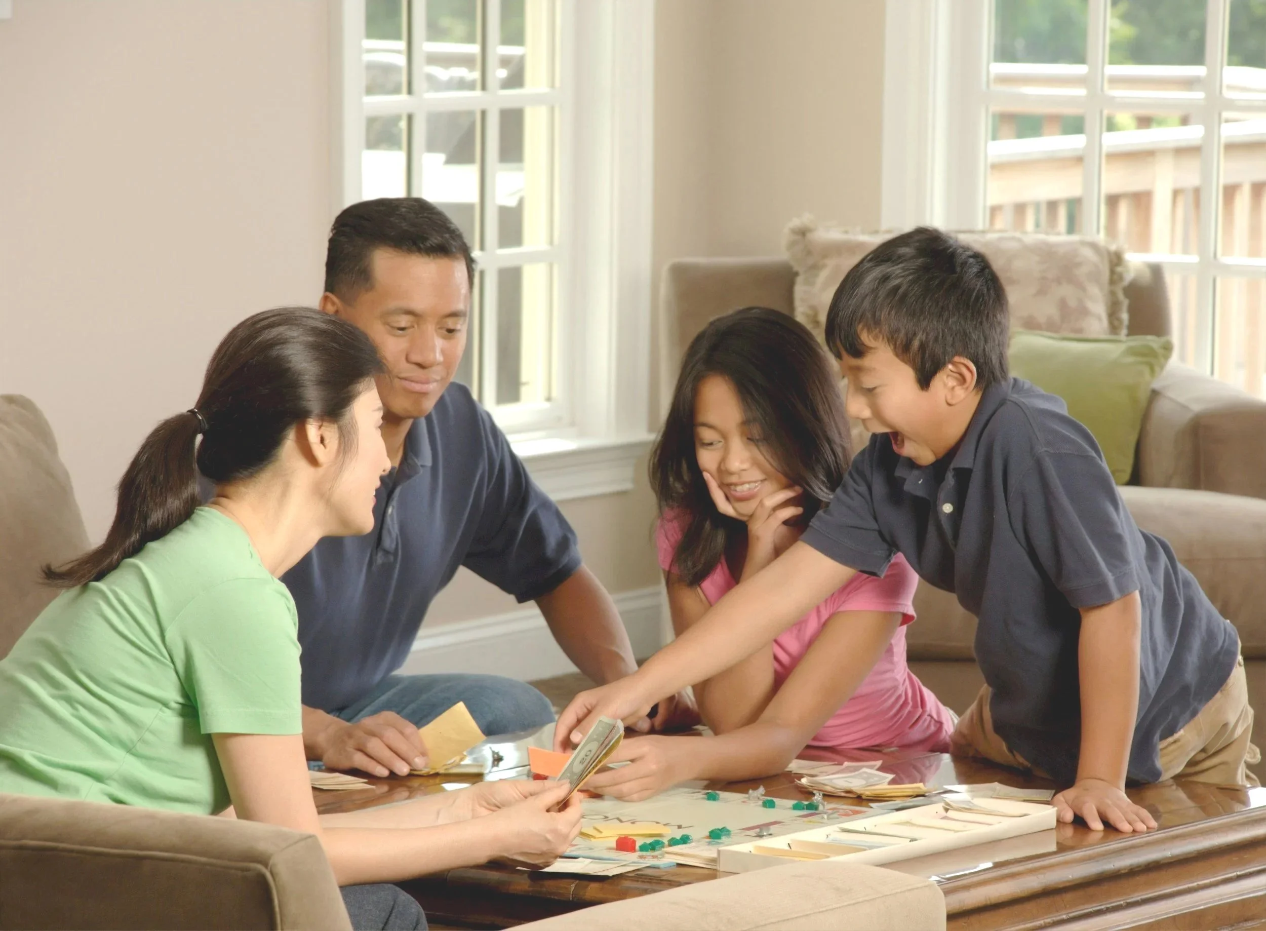 A family of five is gathered around a table playing a board game together in a bright living room with large windows.