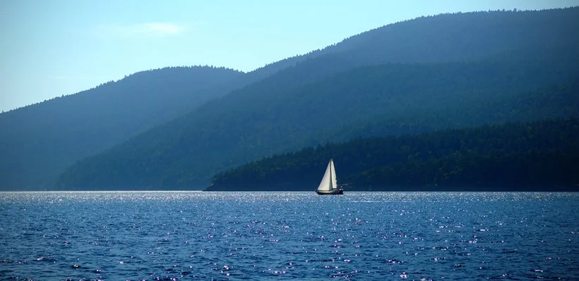 A sailboat on a large body of water with forested hills in the background under a clear sky.