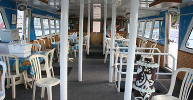 Interior of a boat with tables and chairs, some tables covered with tablecloths, and windows on both sides.