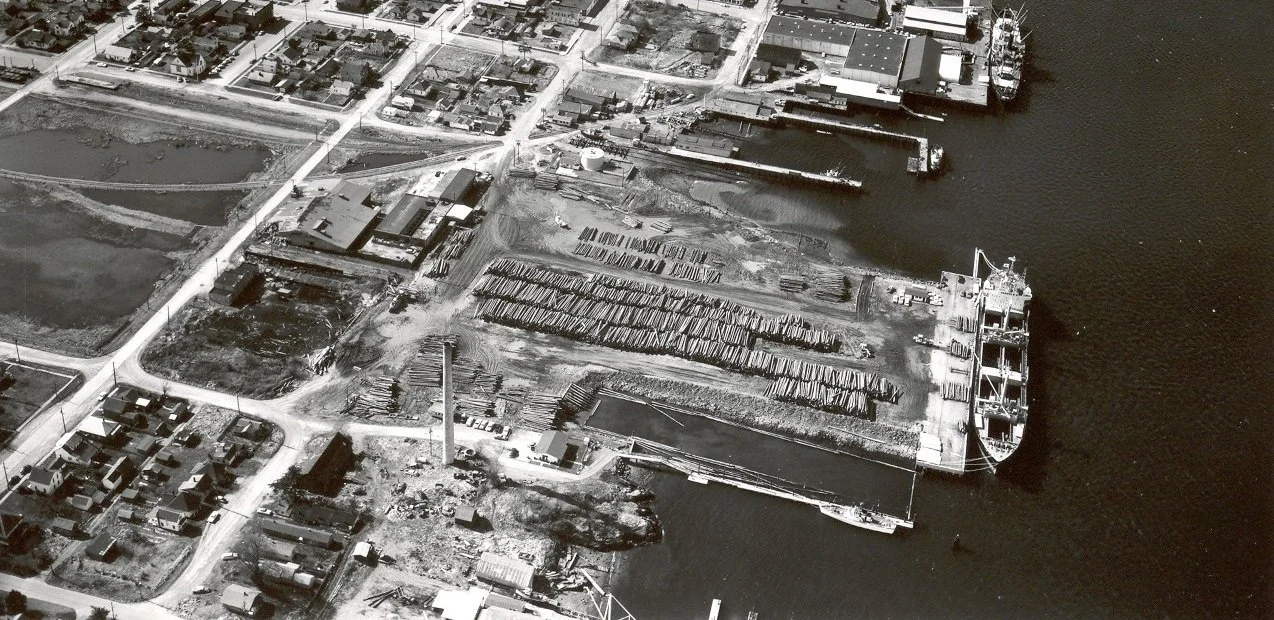 Aerial view of a dockside area with a large cargo ship docked, containing containers and cranes, with surrounding buildings, roads, and residential streets nearby.