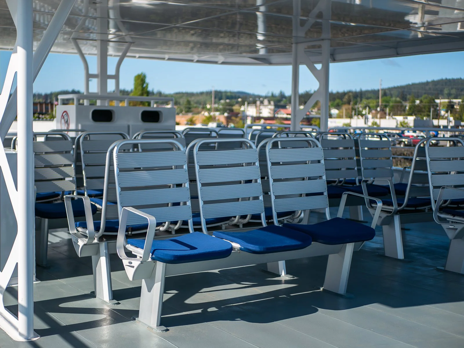 Empty outdoor seating area on a ferry with metal chairs with blue cushions, overlooking a town with hills and buildings in the background.