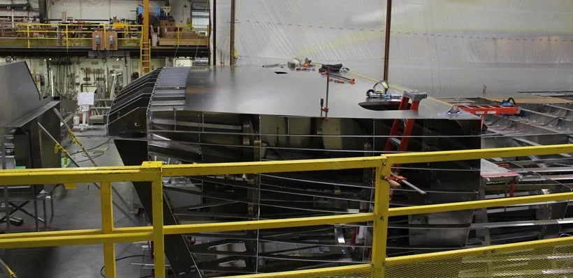 Inside a manufacturing or assembly plant showing partial view of a large black metallic structure, with tools and equipment on top, surrounded by yellow safety railings.