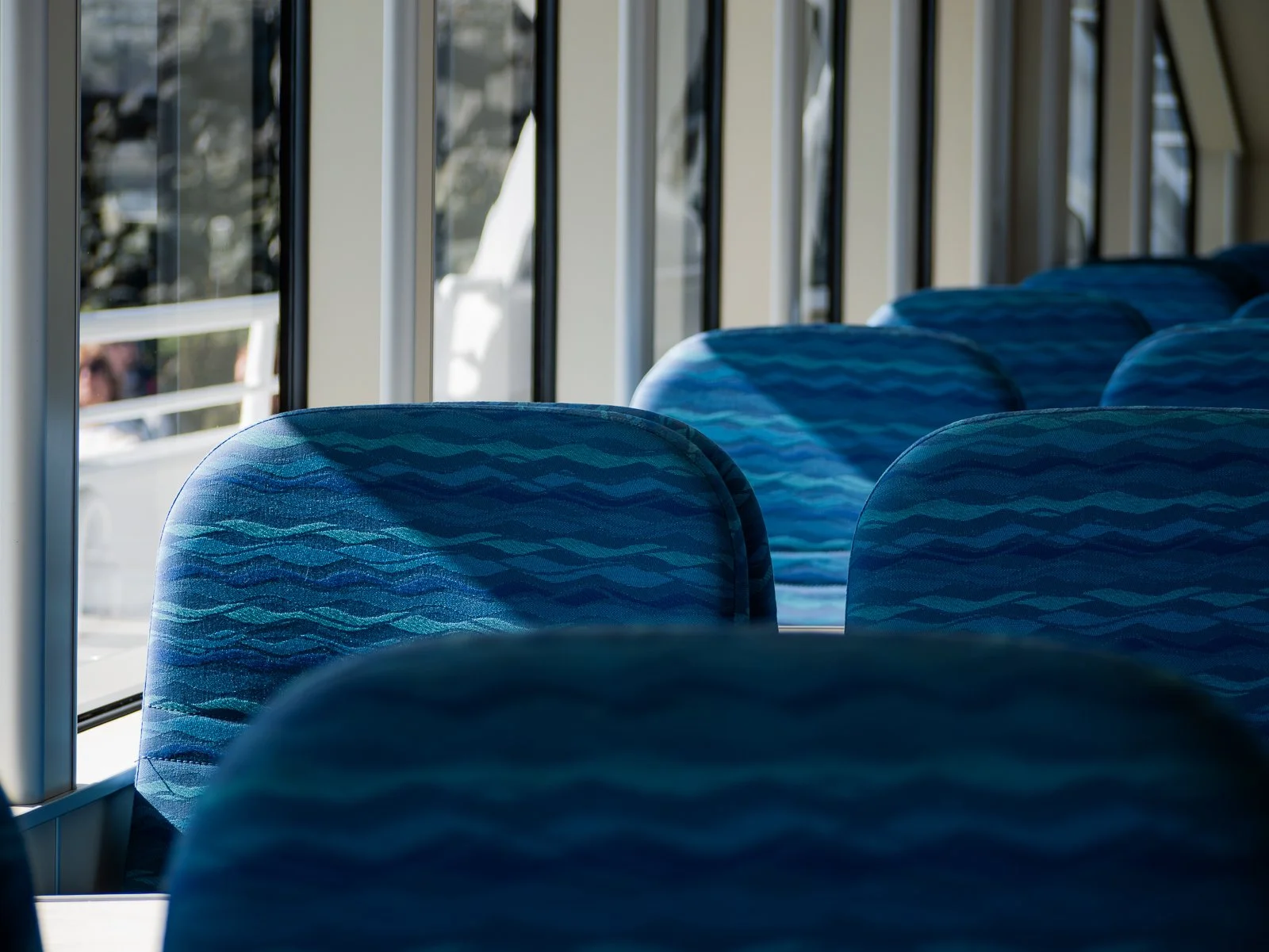 Empty blue patterned bus seats inside a bus with large windows showing sunlight and exterior view.