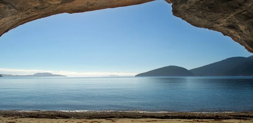 View through a rock arch overlooking calm ocean waters with distant islands and mountain ranges under a clear blue sky.