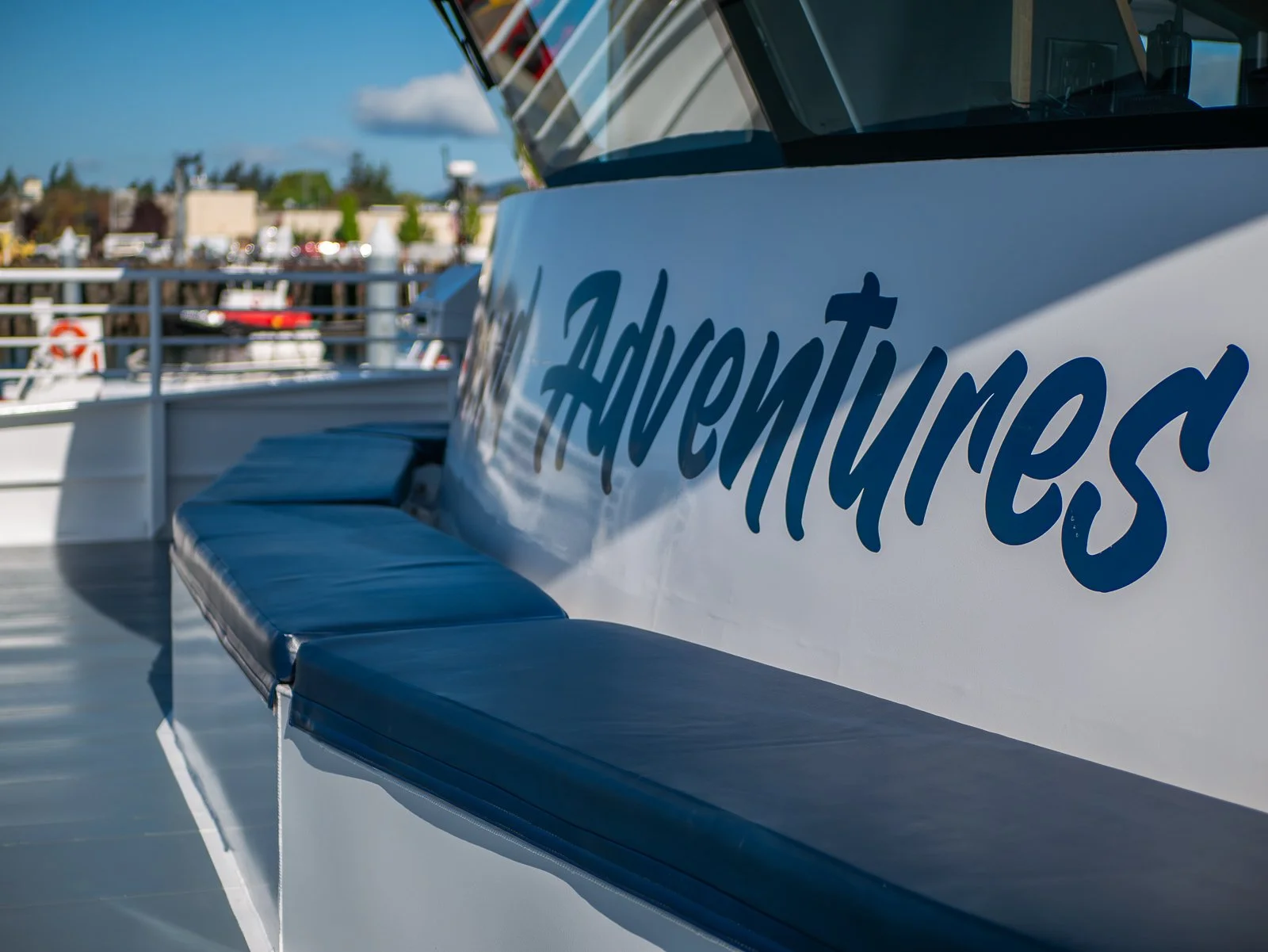 Close-up of a boat with the name 'Adventuress' written on the side, docked at a marina with buildings and boats in the background.