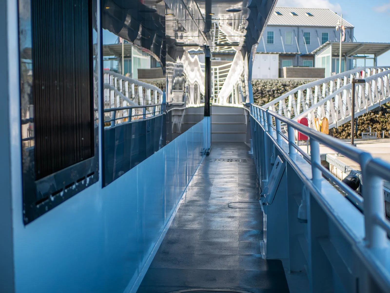 View of a ferry boat walkway with reflective metallic sides, handrails, and stairs leading outside, with buildings in the background.
