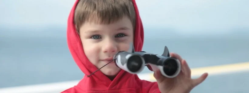 A young boy in a red hoodie holds binoculars up to his eyes on a boat or dock, with water in the background.