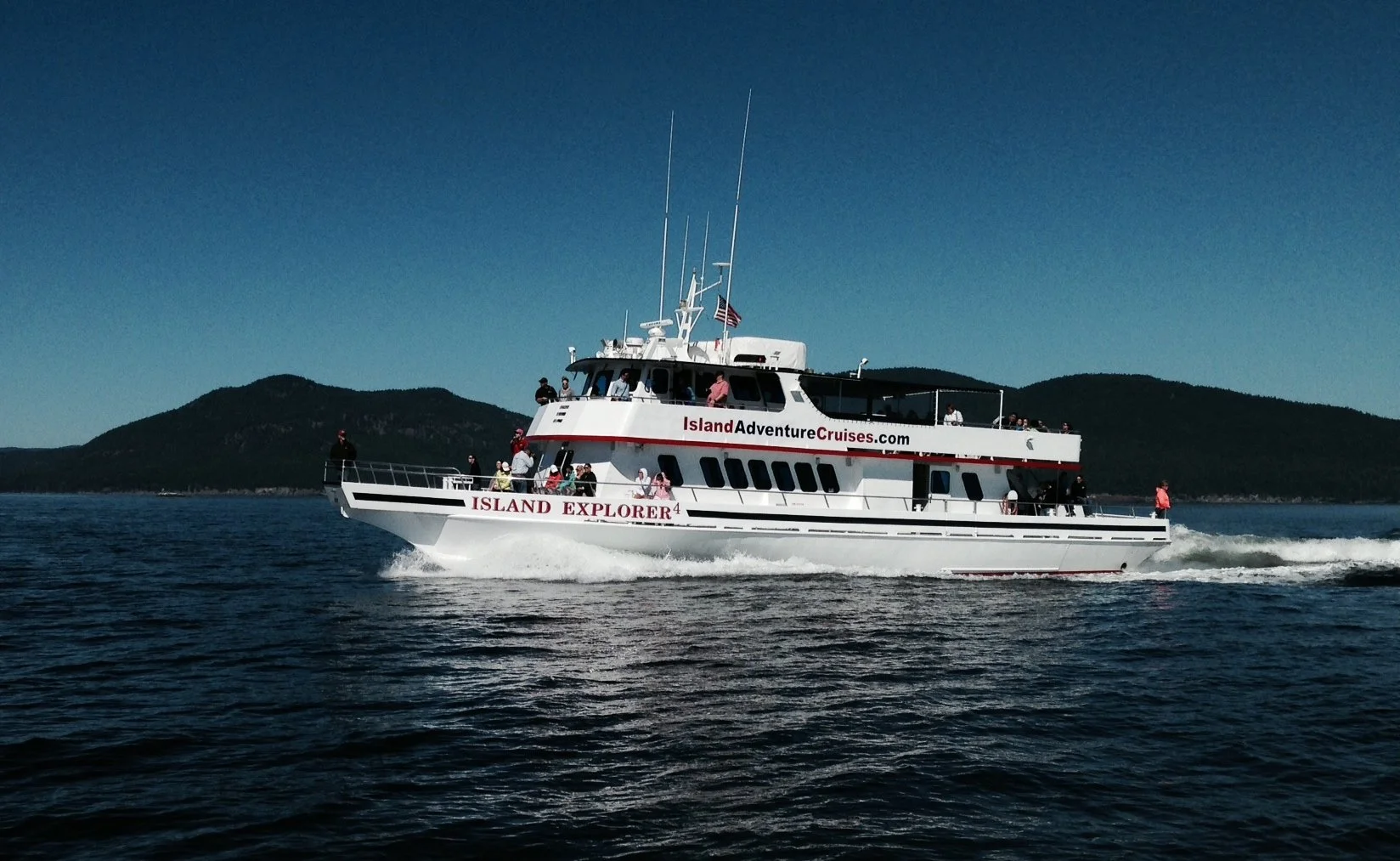 A white island cruise boat named Island Explorer on the water with a dark mountain landscape in the background, carrying passengers on a daytime cruise.