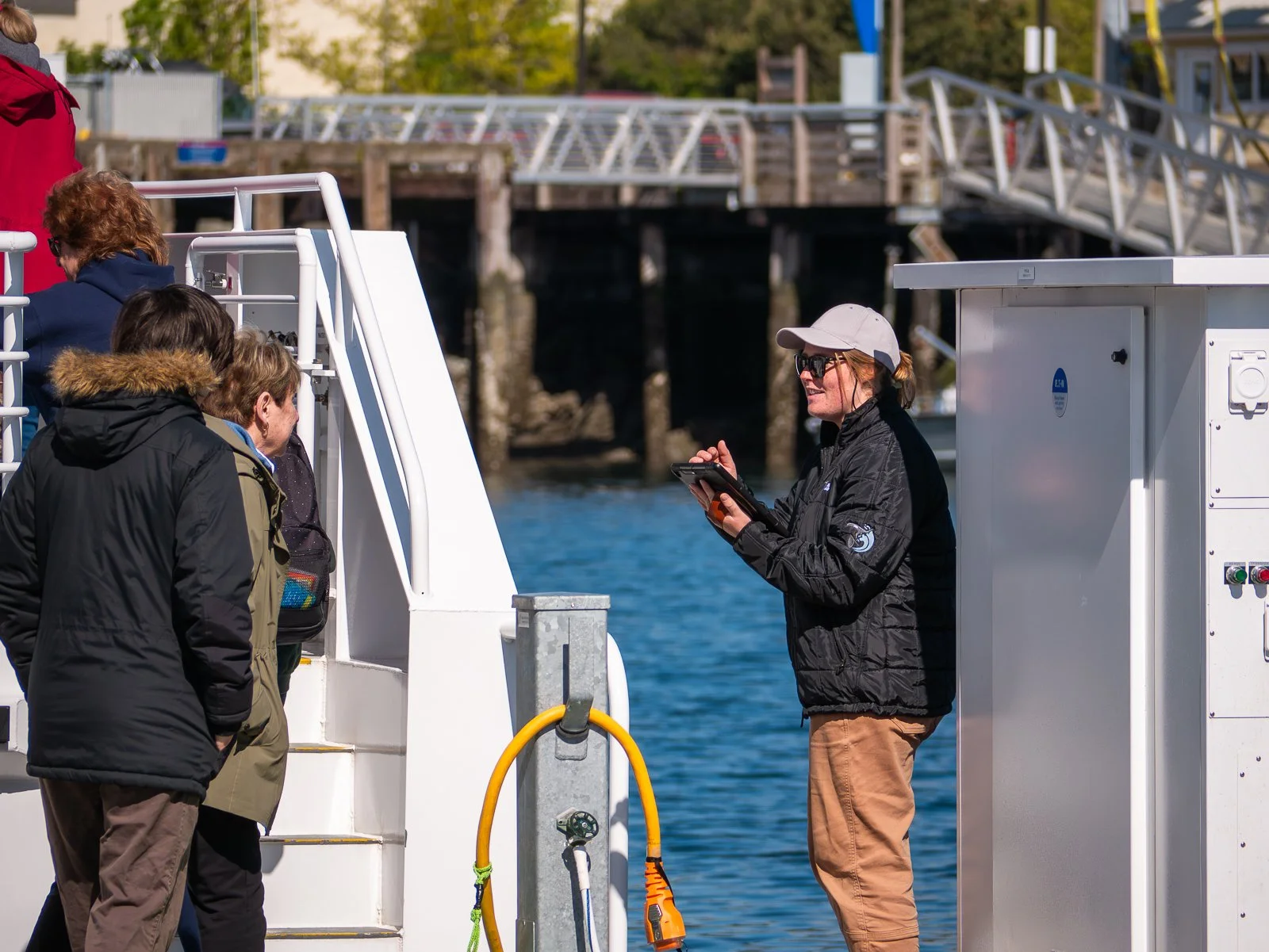 Group of people boarding a boat at dock, woman with tablet assisting, water and dock structure in background.
