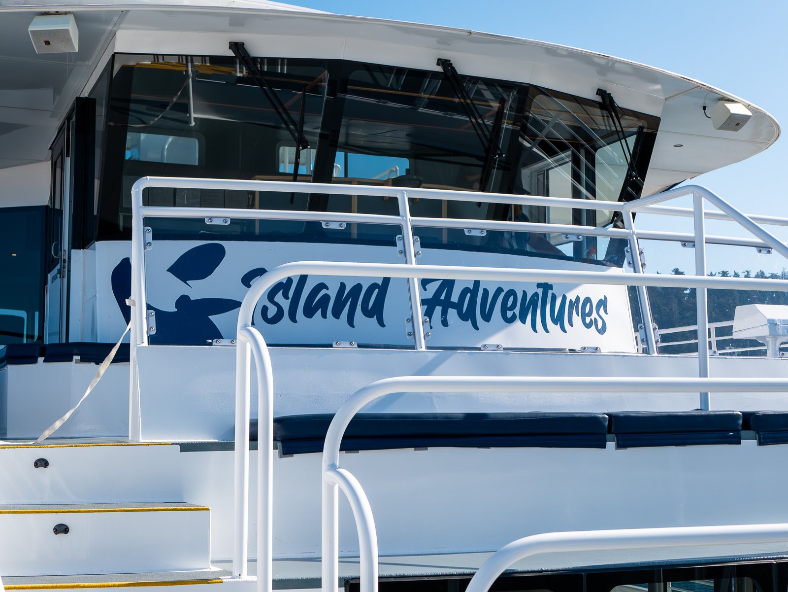 Close-up of a boat with the words 'Island Adventures' written on its side, featuring a railing and pool area, under a clear blue sky.