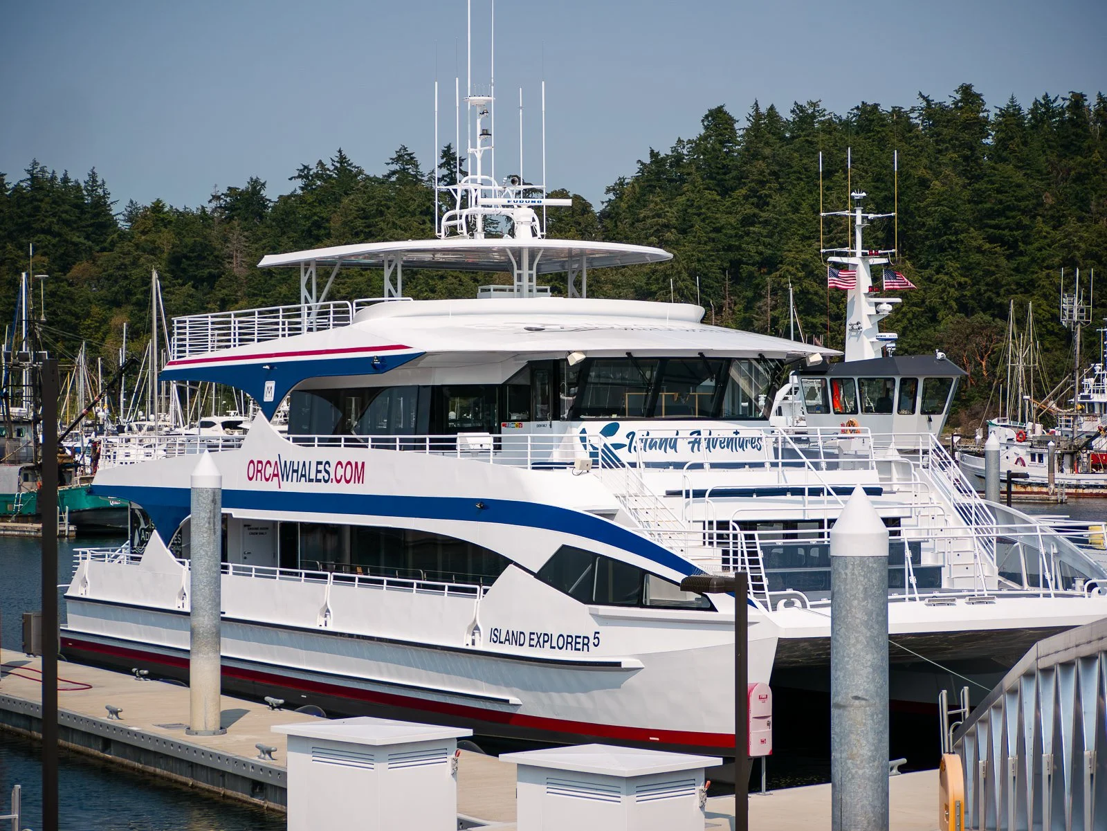 A large white and blue yacht named Island Explorer 5 docked at a marina, with trees in the background and several other boats nearby.