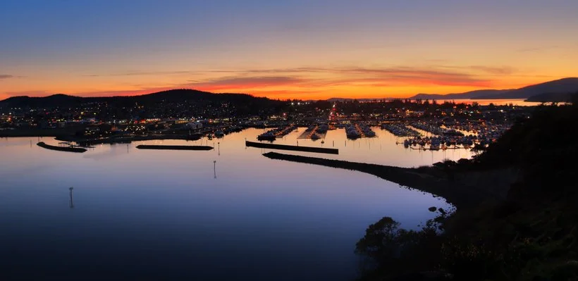 Sunset over a harbor with boats docked, city lights beginning to turn on, mountains in the background, calm water reflecting the sky.