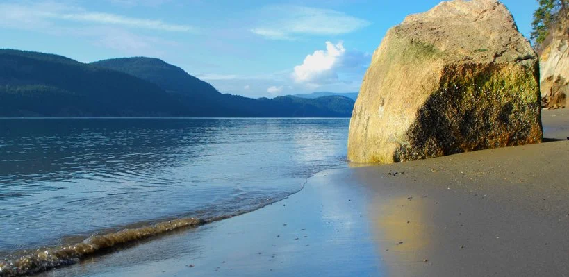 A peaceful beach scene with a large rock on the sandy shore, calm waters, distant mountains, and a bright blue sky with some clouds.