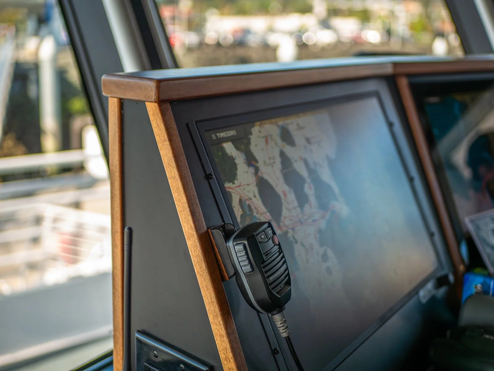 Close-up of a navigation screen on a boat with a mounted radio or communication device, framed by wooden paneling, with blurred outdoor marina in the background.